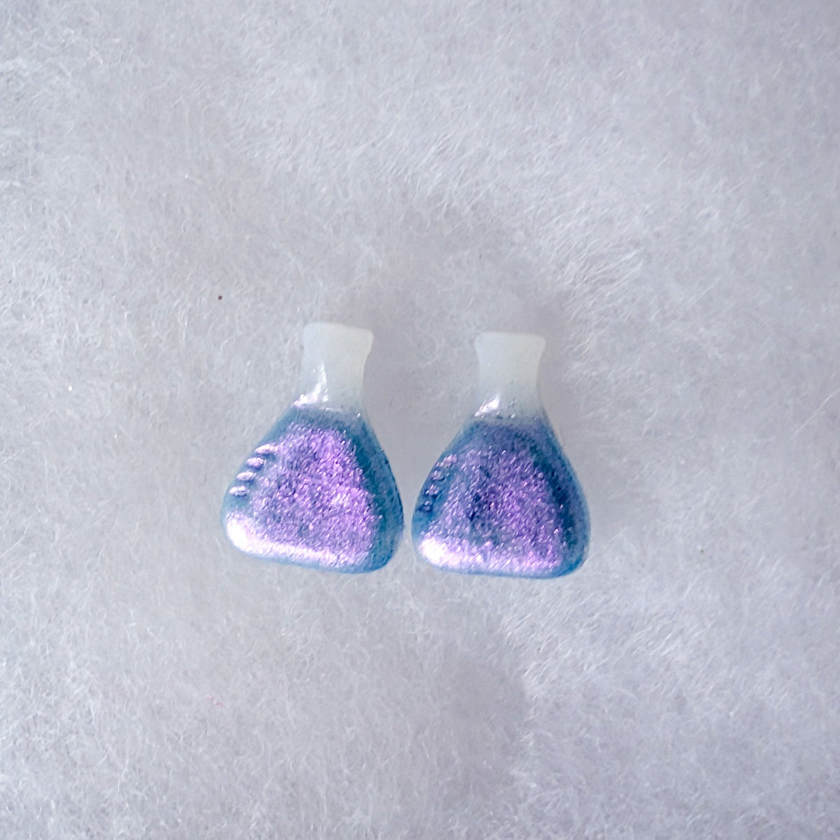 A pair of beaker shaped stud earrings with a white top and a glittery purple-to-blue colorshift finish.