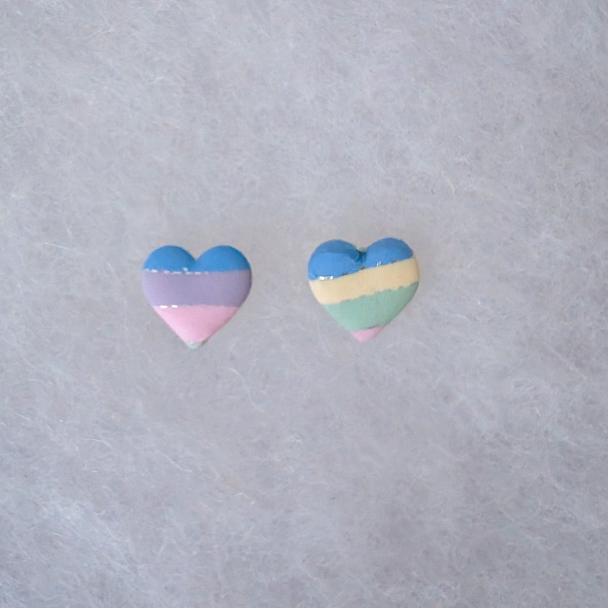Two small heart-shaped stud earrings with horizontal pastel stripes in blue, lavender, pink on one earring and blue, cream, mint, and pink on the other with silver accents.