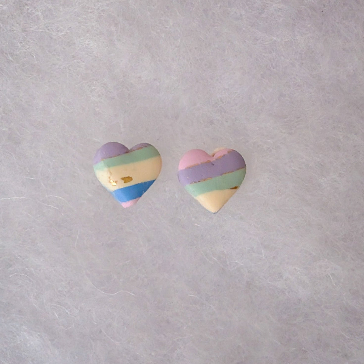 Two small heart-shaped earrings with pastel rainbow stripes in purple, blue, green, yellow, and pink with gold accents.
