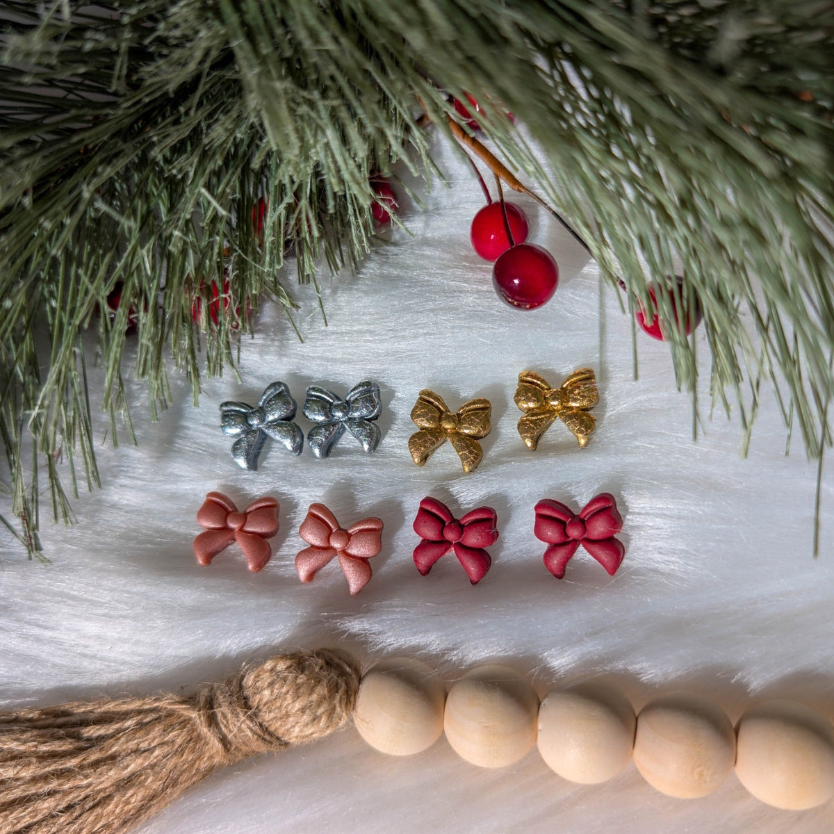 Four colors of coquette bow stud earrings arranged beneath snowy evergreen branches