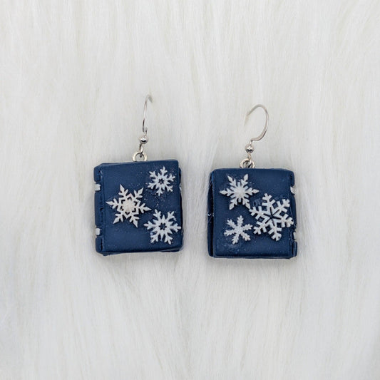 Navy blue miniature book earrings decorated with white snowflakes and silver-tone hooks.