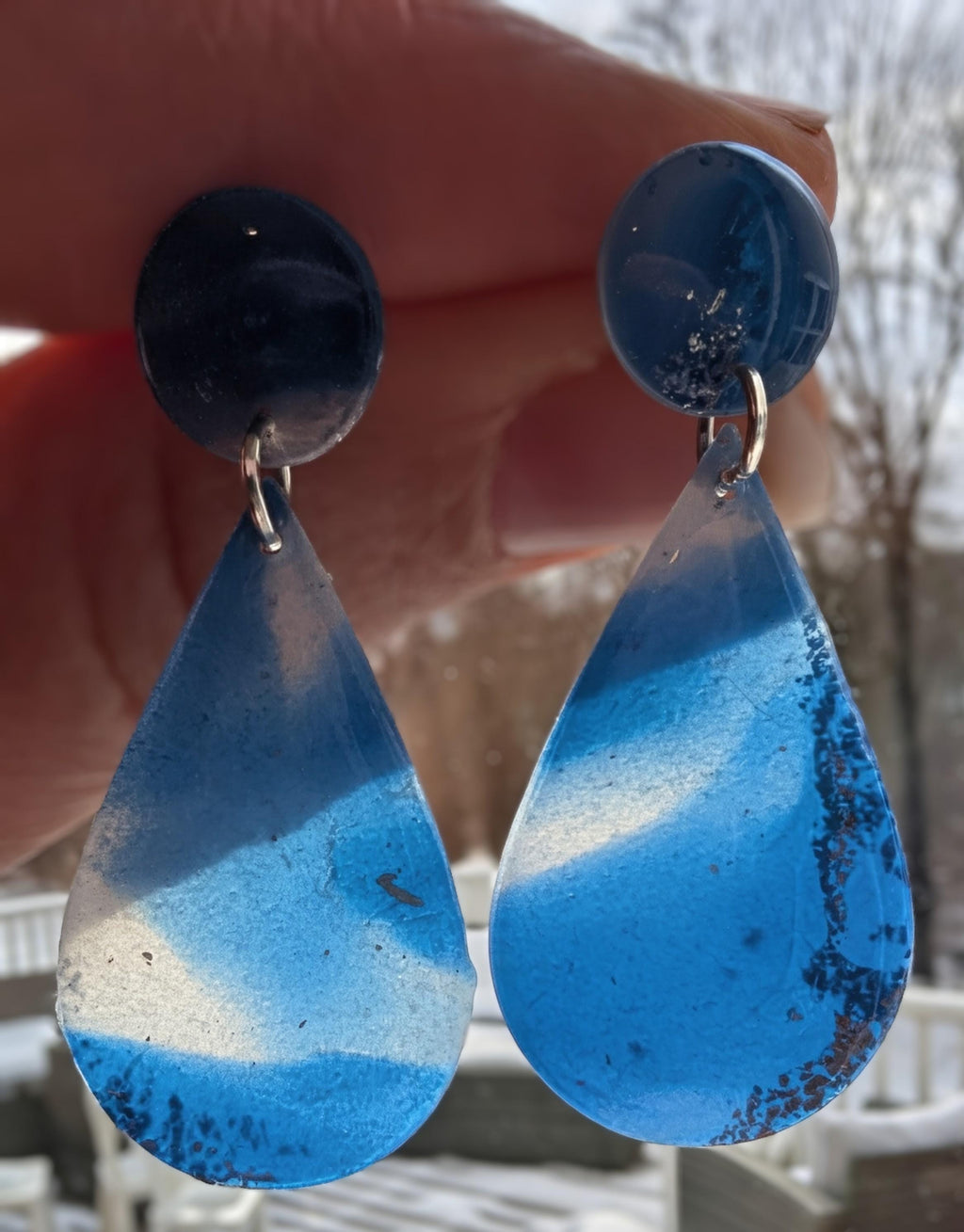Teardrop-shaped blue earrings held by a hand with a blurred outdoor background.