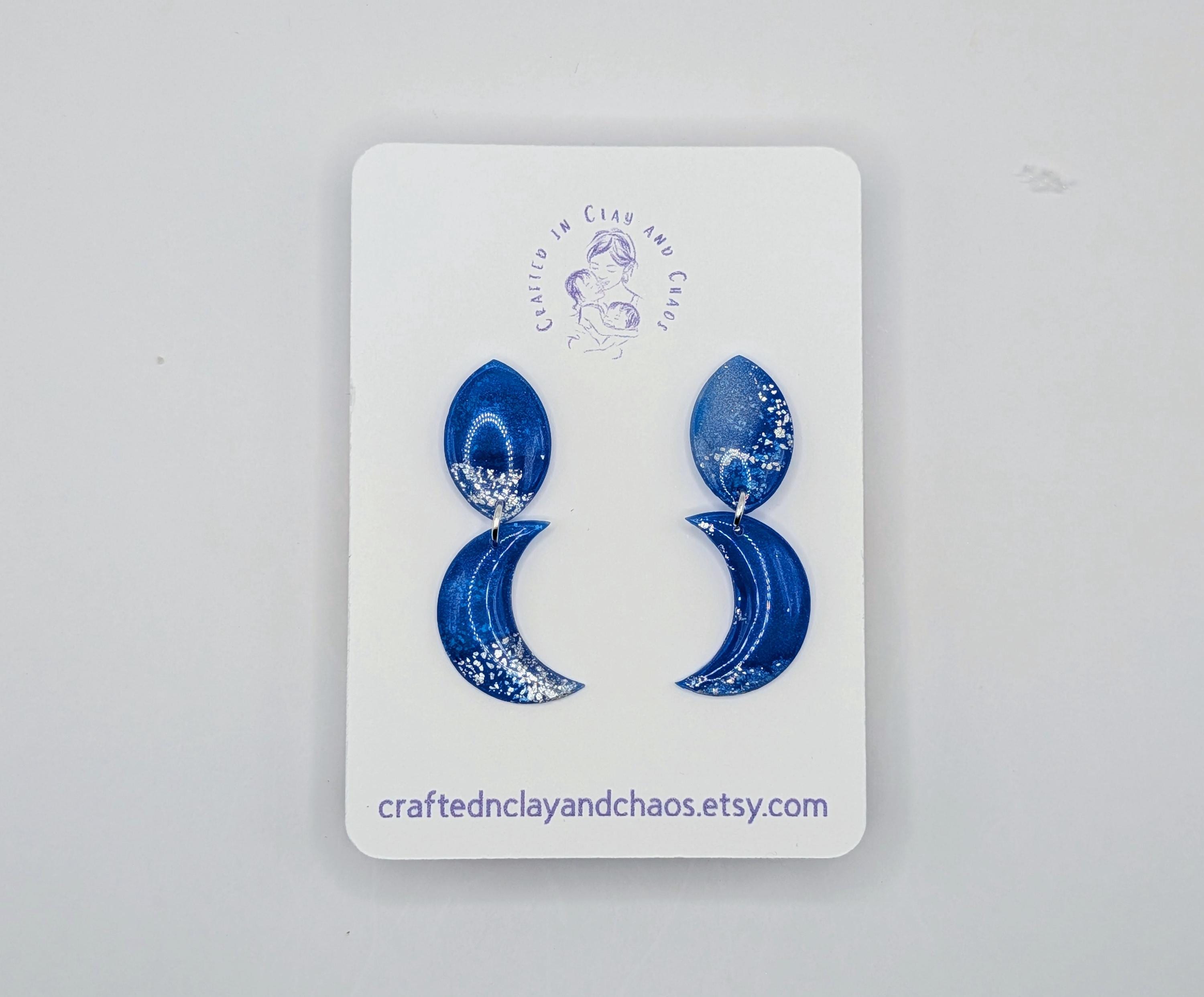 Blue crescent moon earrings on a white card with a brand logo and website text.