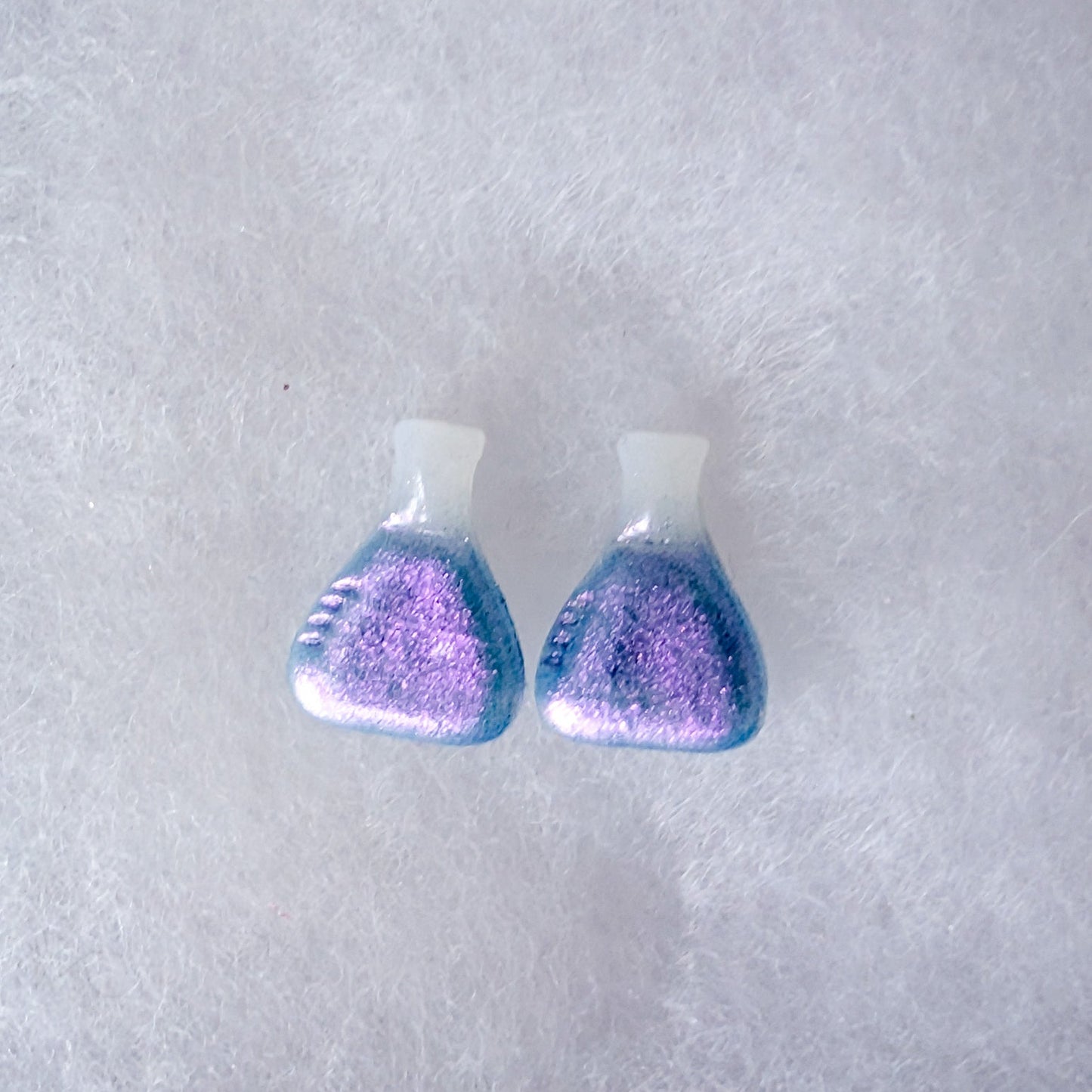 A pair of beaker shaped stud earrings with a white top and a glittery purple-to-blue colorshift finish.