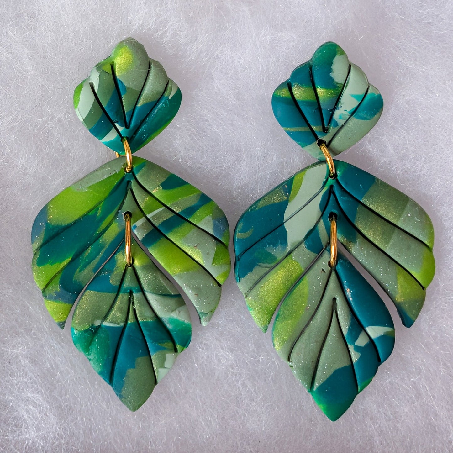 A pair of handmade polymer clay dangle earrings shaped like layered leaves, featuring a marbled pattern of green, teal, and blue with small gold metal hooks, displayed on a soft white textured background.