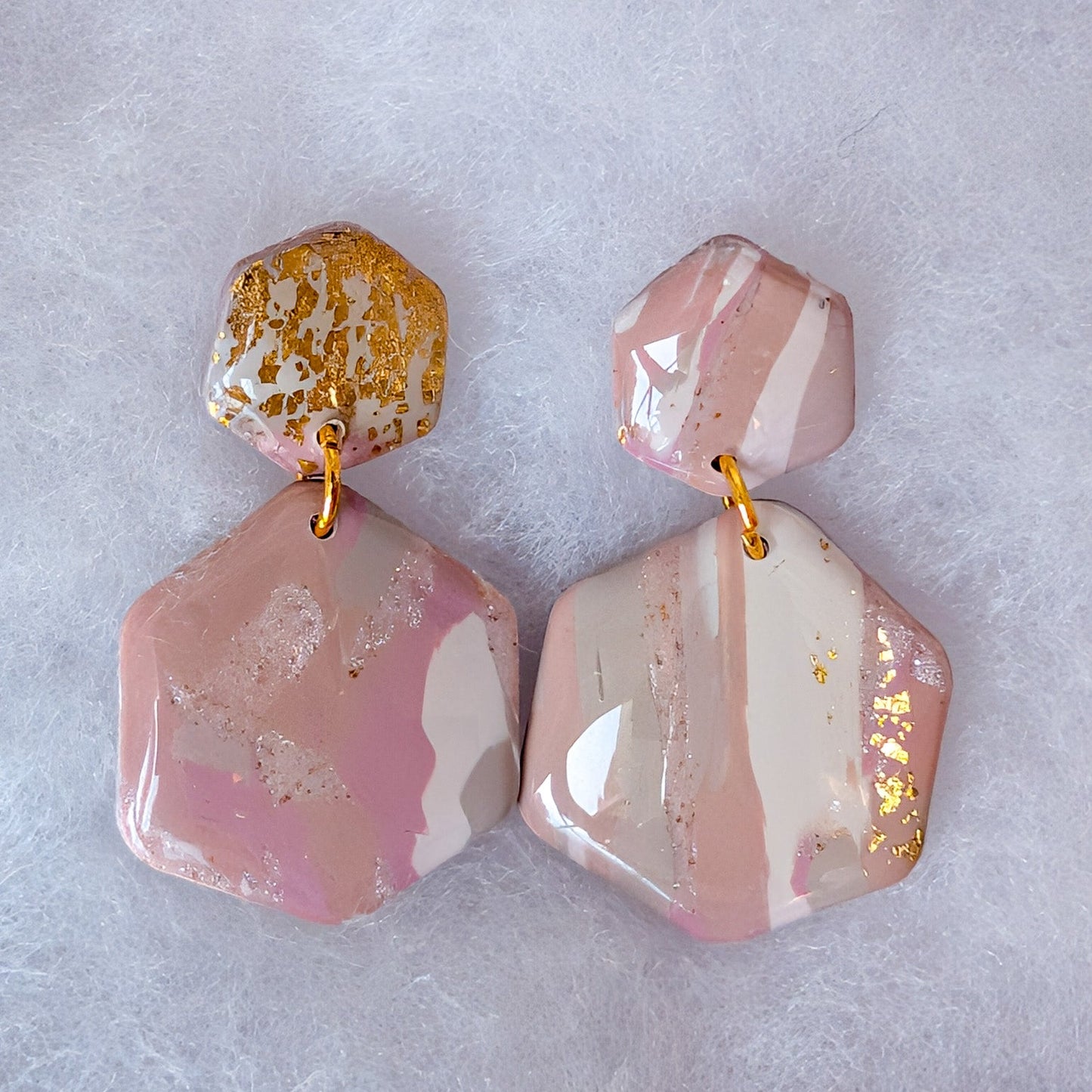 A pair of chunky, geometric drop earrings made from pink and white marbled stone with scattered gold leaf flakes, connected by small gold rings, displayed on a textured white surface.