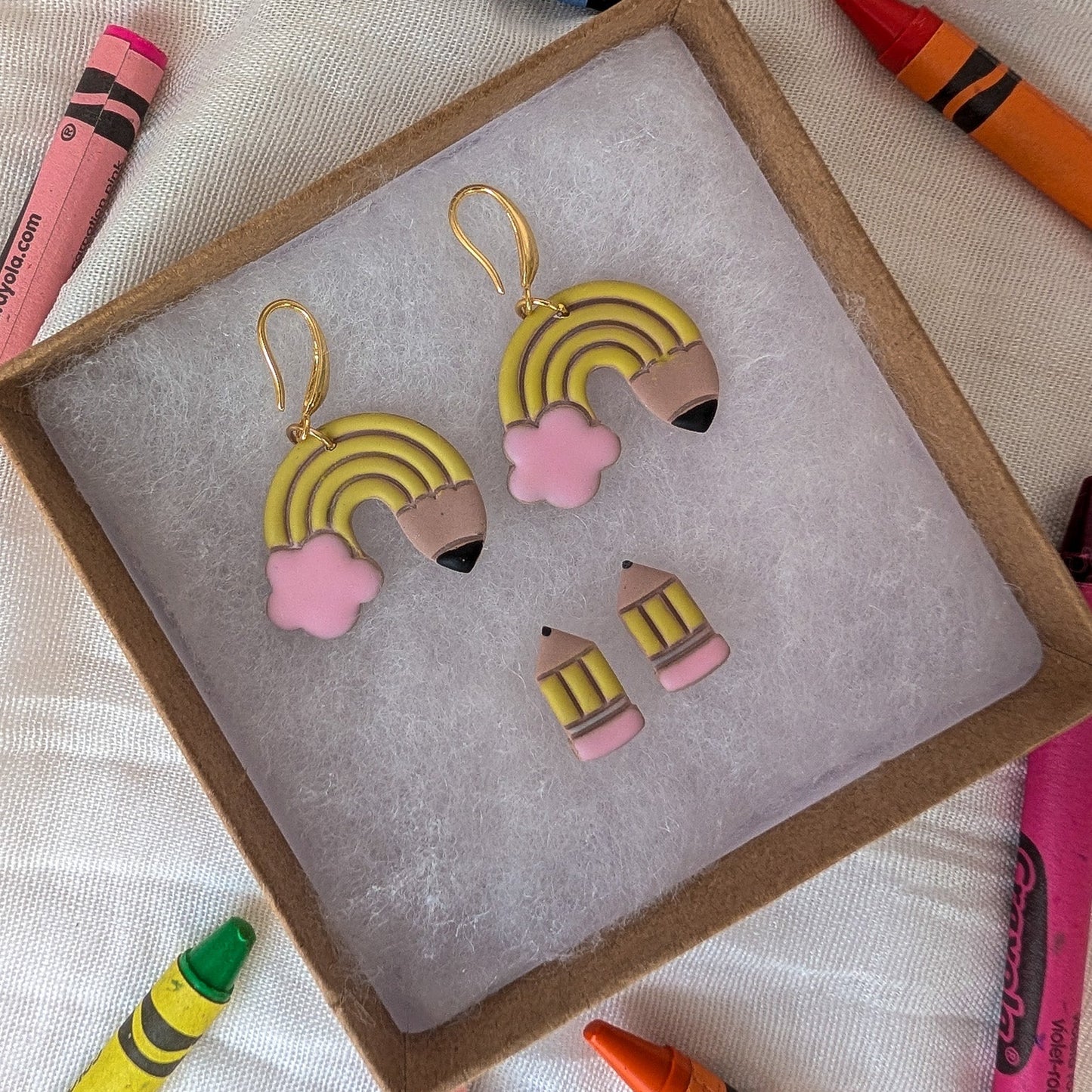 A pair of rainbow pencil earrings displayed in a brown gift box with a gray insert, surrounded by colorful crayons on a white fabric background.