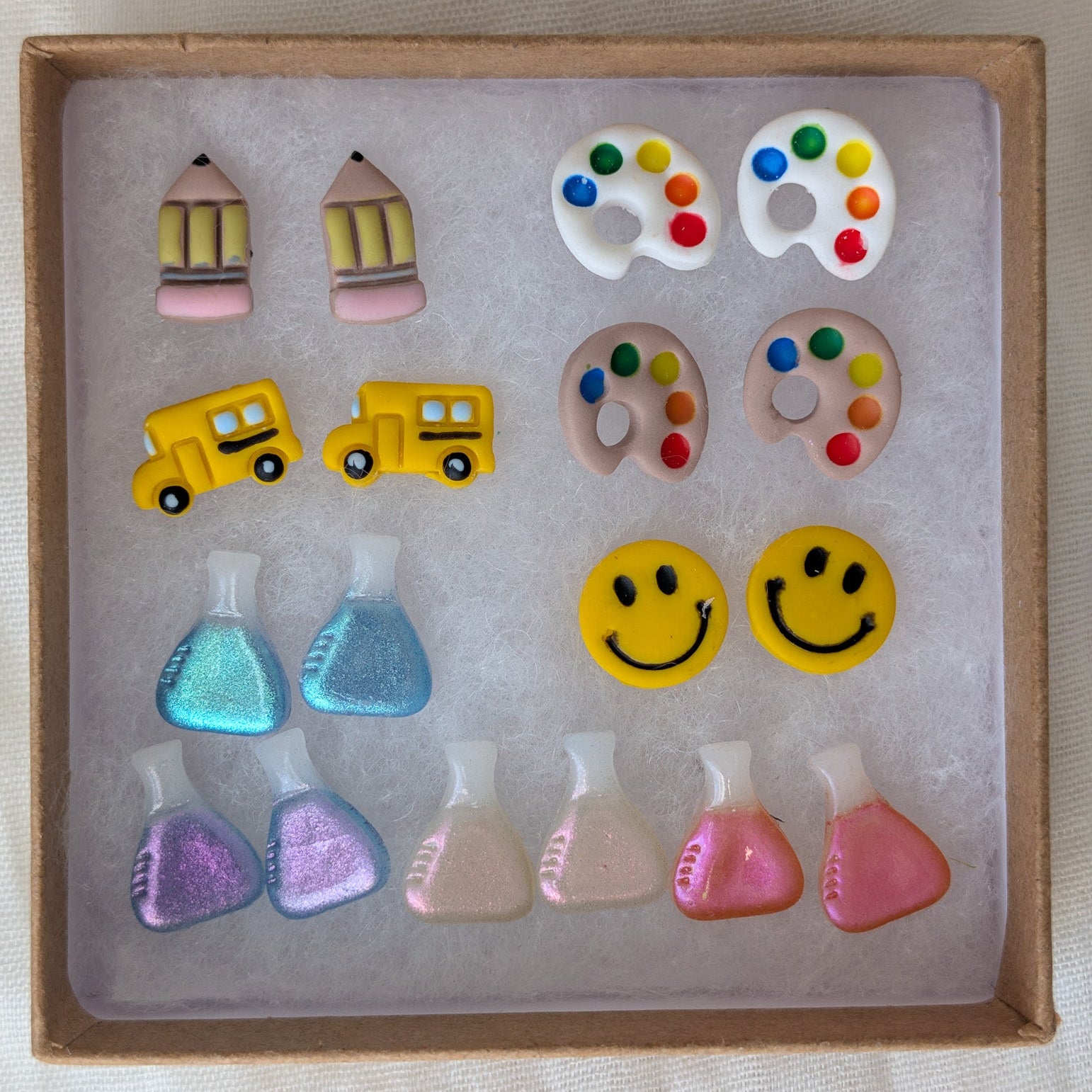 A set of colorful novelty earrings displayed inside a square cardboard box on a white textured fabric background. The earrings include pencils, paint palettes, school buses, smiley faces, and small colorful bottle-shaped charms.