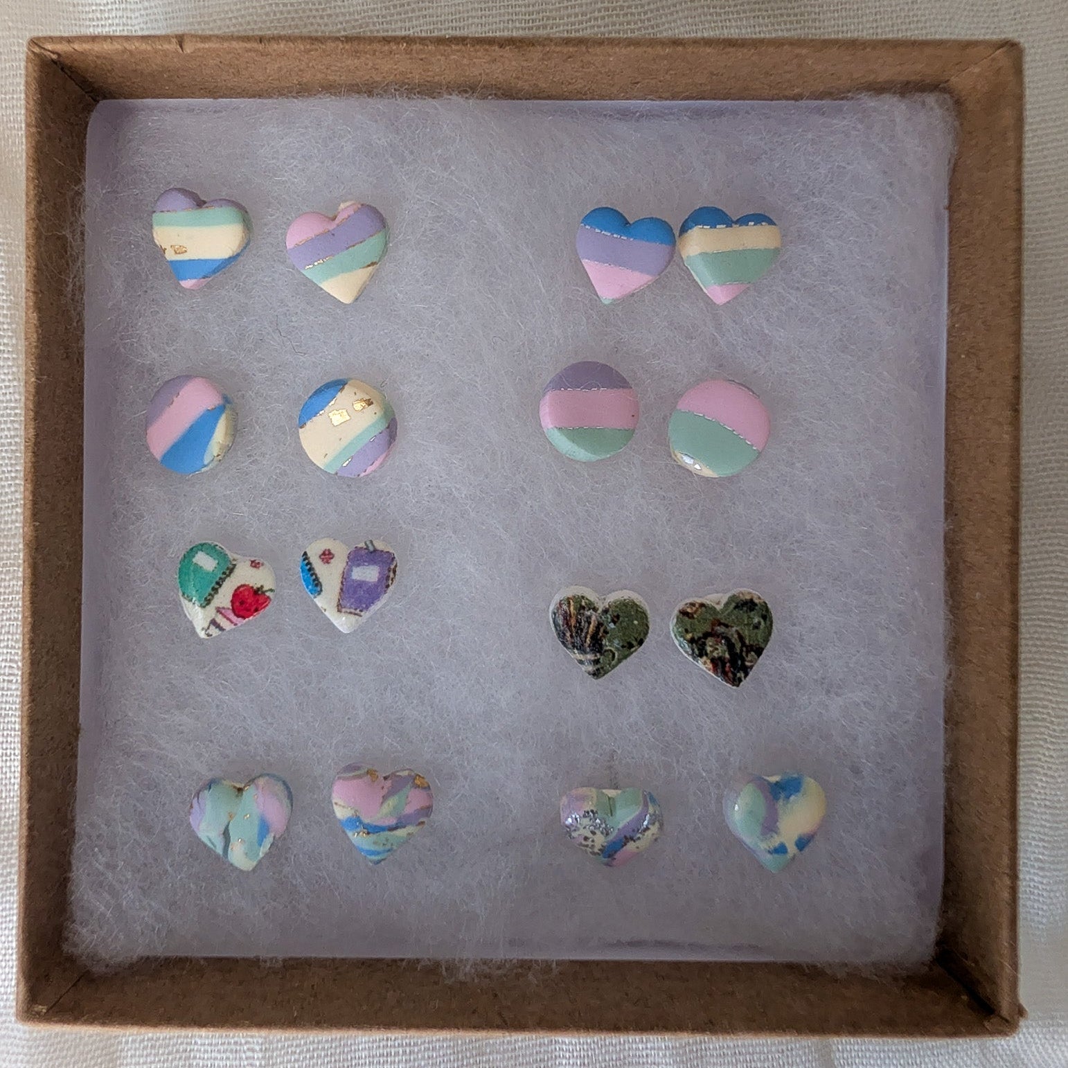 A set of small heart-shaped stud earrings displayed inside a brown cardboard box lined with light gray tissue paper. The earrings feature pastel rainbow stripes, marble patterns, and one pair with a green-and-black marbled design.