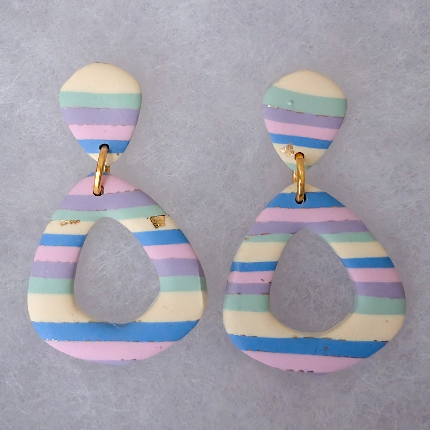 A pair of handmade hollow diamond taerdrop shaped dangle earrings with horizontal pastel stripes in pink, blue, purple, green, and cream, suspended by gold-colored rings.