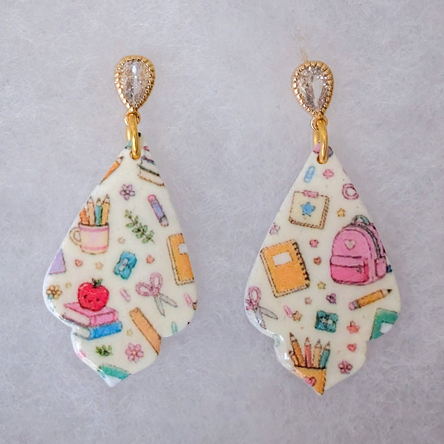 A pair of teardrop-shaped drop earrings with a white background and a colorful school-themed pattern featuring backpacks, notebooks, pencils, and books, hanging from gold-toned posts.