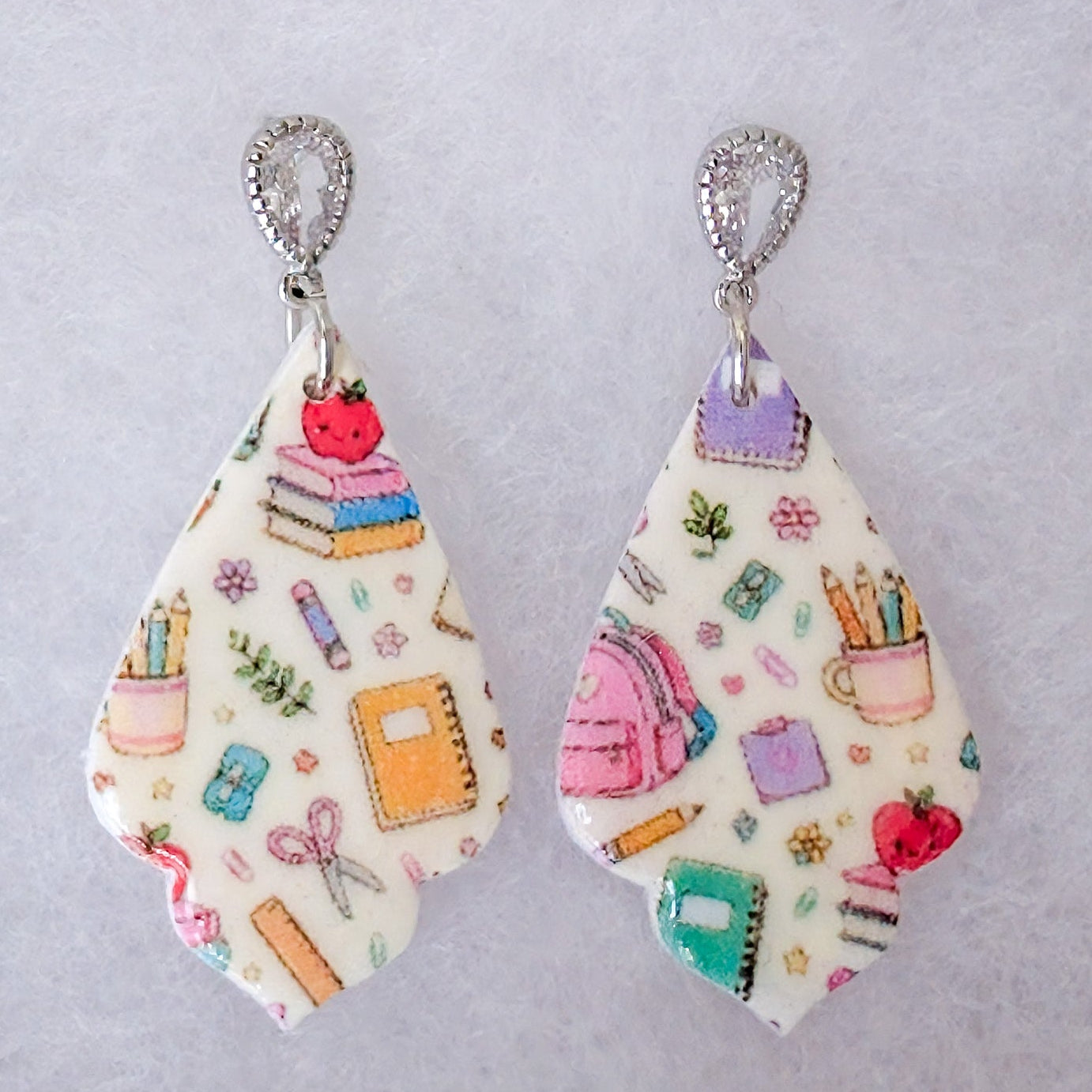 A pair of teardrop-shaped dangle earrings with a white background and a colorful school supply pattern, including books, pencils, a backpack, and an apple, hanging from silver hooks.