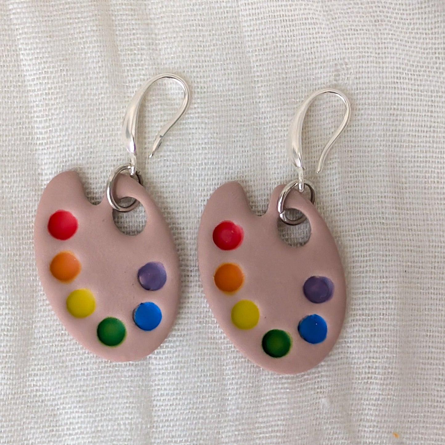 A pair of dangling earrings shaped like pastel paint palettes with colorful paint droplets and silver fishhook wires.