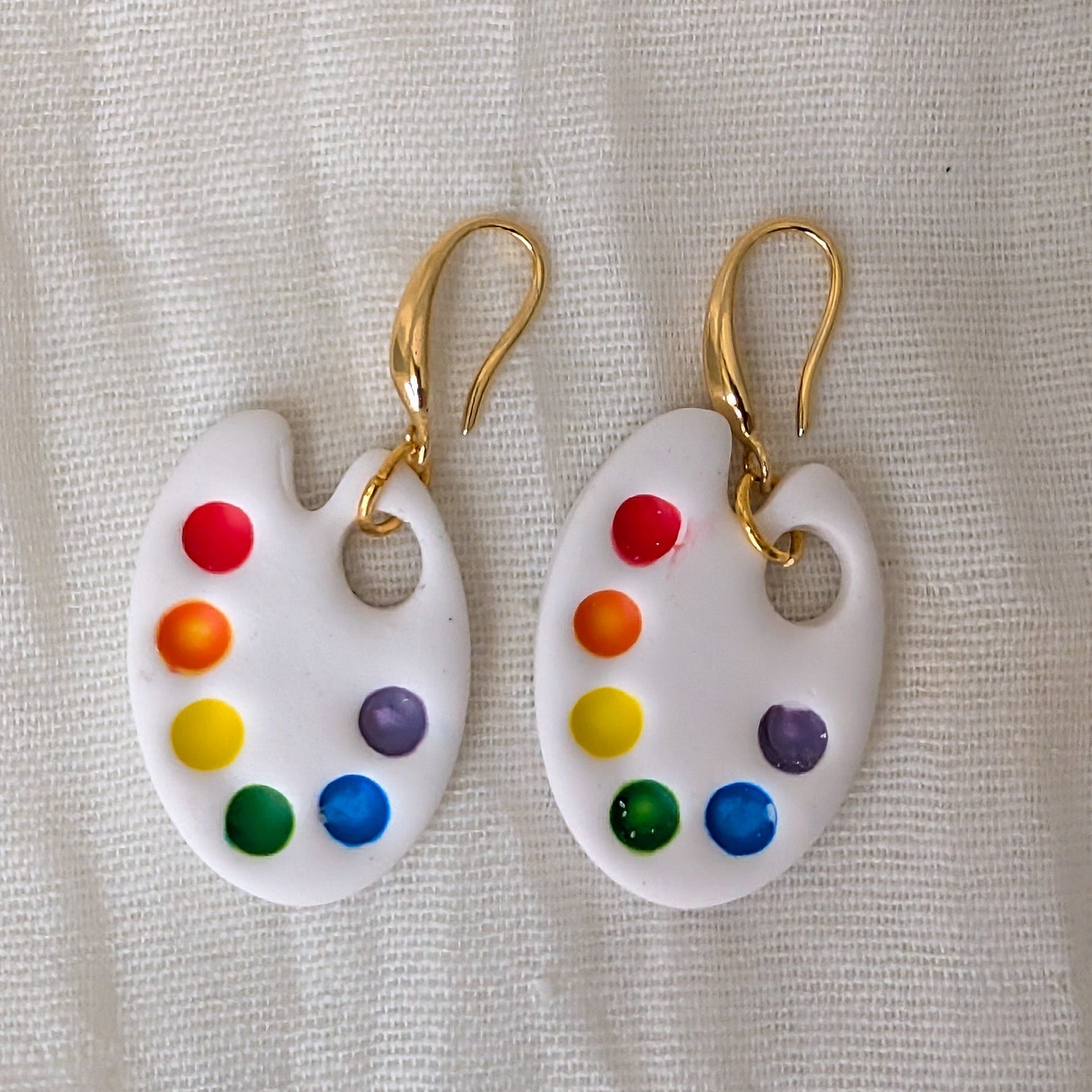 A pair of white polymer clay earrings shaped like an artist's paint palette, featuring colorful painted paint droplets in red, orange, yellow, green, blue, and purple, hanging from 24K gold-tone fishhook ear wires.