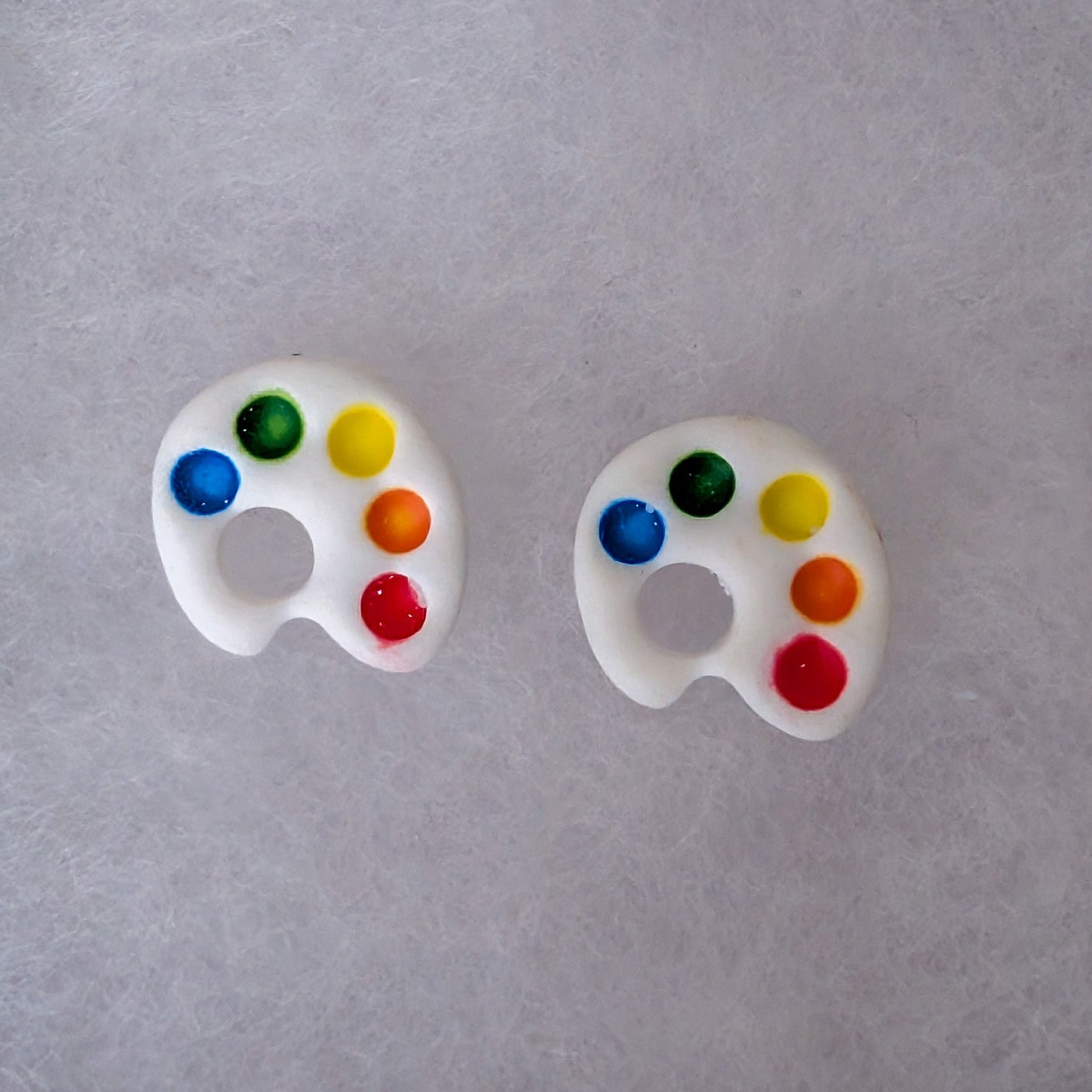 A pair of white stud earrings shaped like miniature paint palettes, featuring colorful faux paint wells in blue, green, yellow, orange, and red.