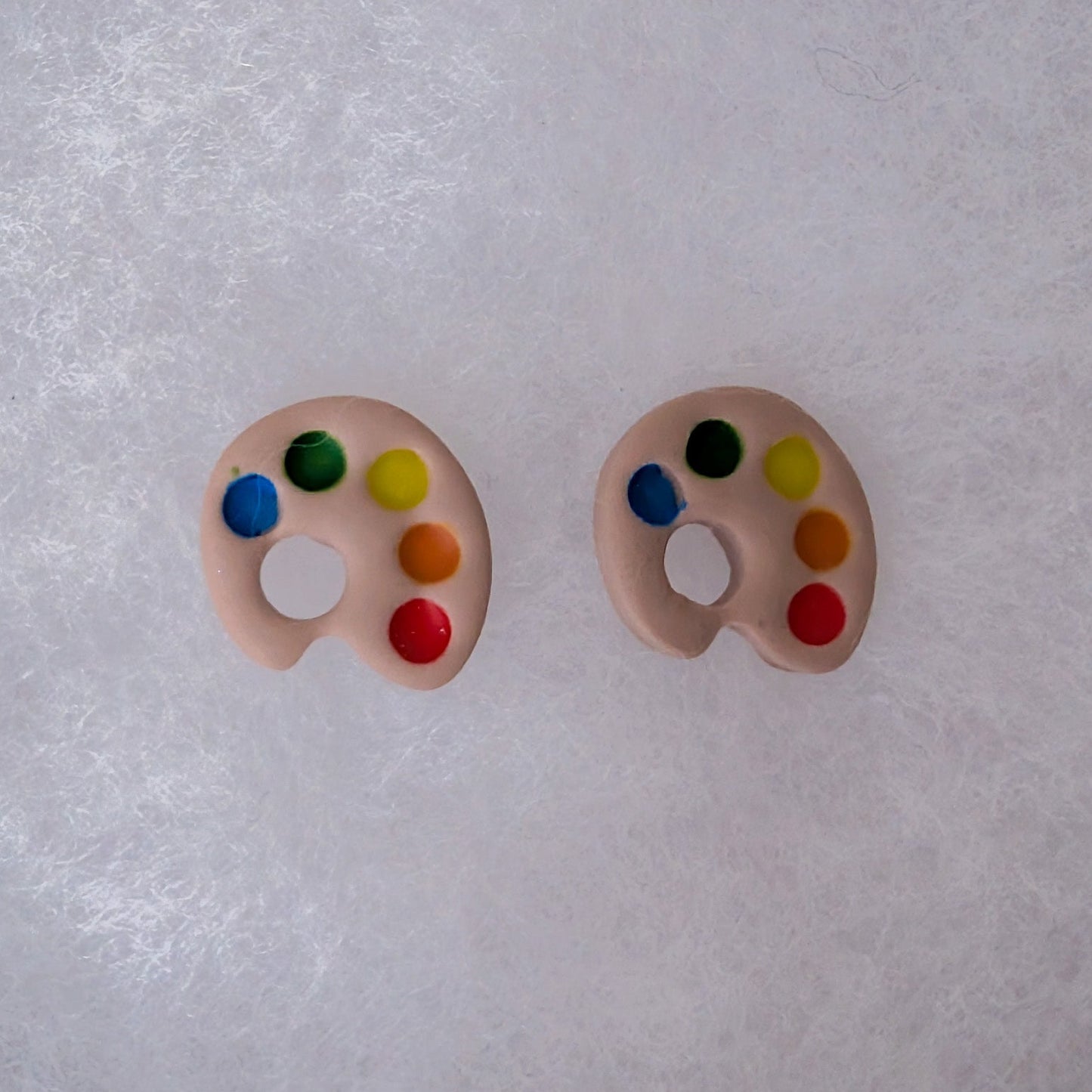 A pair of clay earrings shaped like miniature artist paint palettes, featuring a beige base with colorful dots representing paint colors.