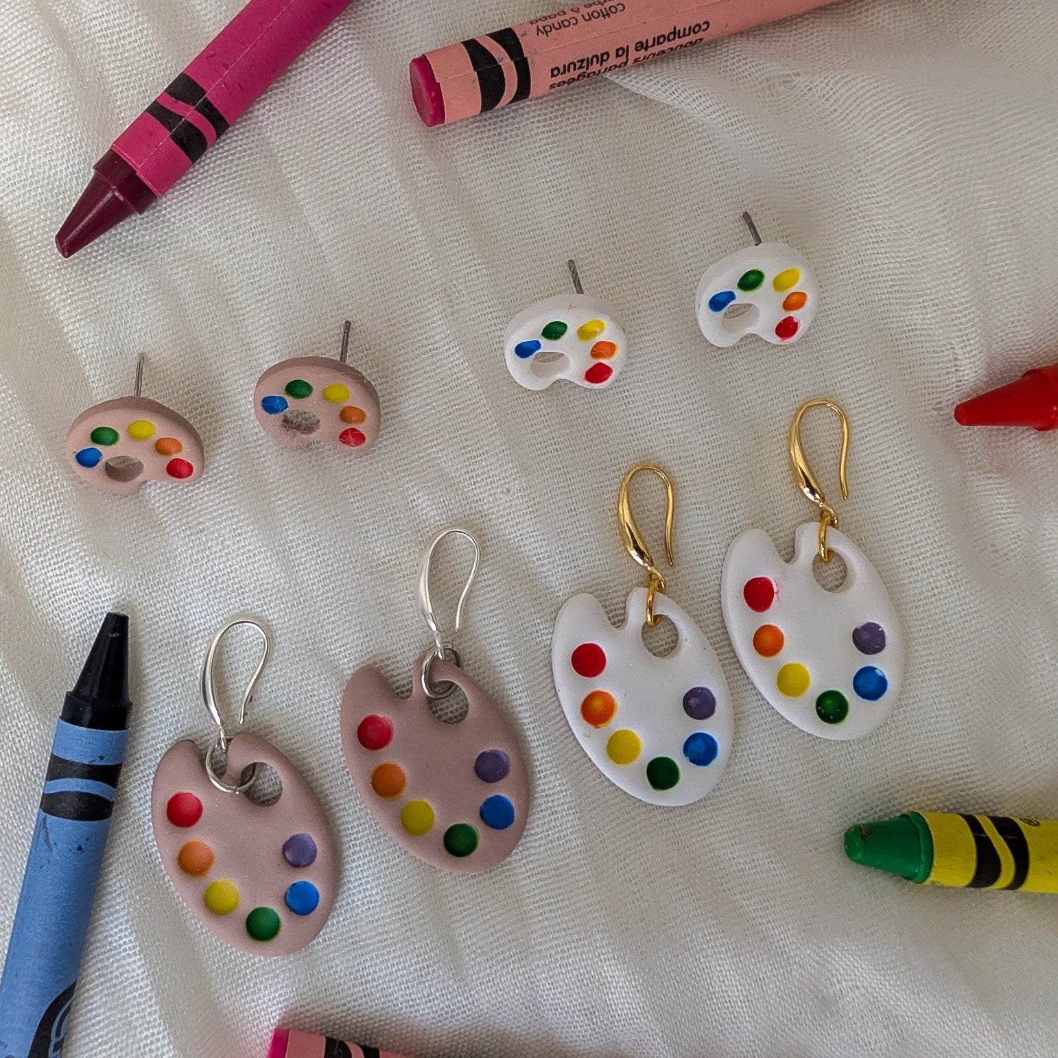 A styled flat-lay of small artist-themed jewelry pieces on a white fabric background, including several white and pink paint palette charms with colorful paint-dot details and matching small earrings, surrounded by crayons.