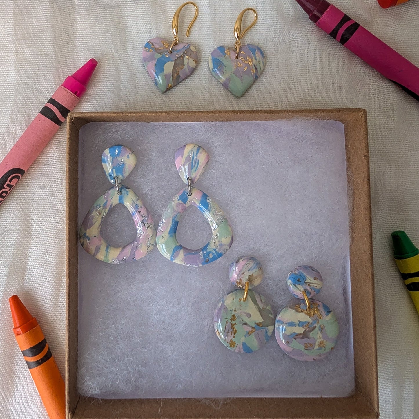 Three pairs of handmade marbled resin drop earrings with gold hooks are displayed on a white fabric background, surrounded by colorful crayons. One pair is shown outside a square cardboard tray, while the other two pairs are inside the tray.