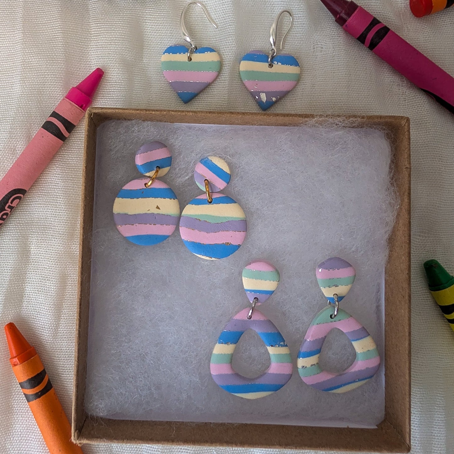 A set of handmade pastel striped earrings displayed in a small brown box on white tissue paper, surrounded by colorful crayons on a white fabric background.