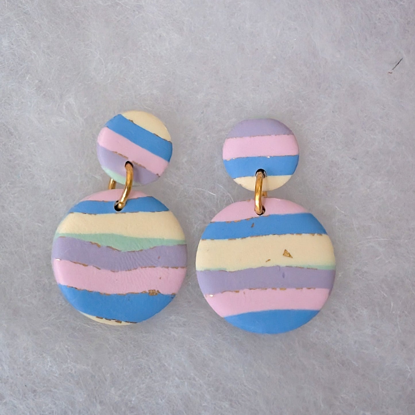 A pair of handmade dangle earrings with circular disc charms featuring pastel horizontal stripes in blue, pink, purple, and cream, suspended from gold-tone rings.