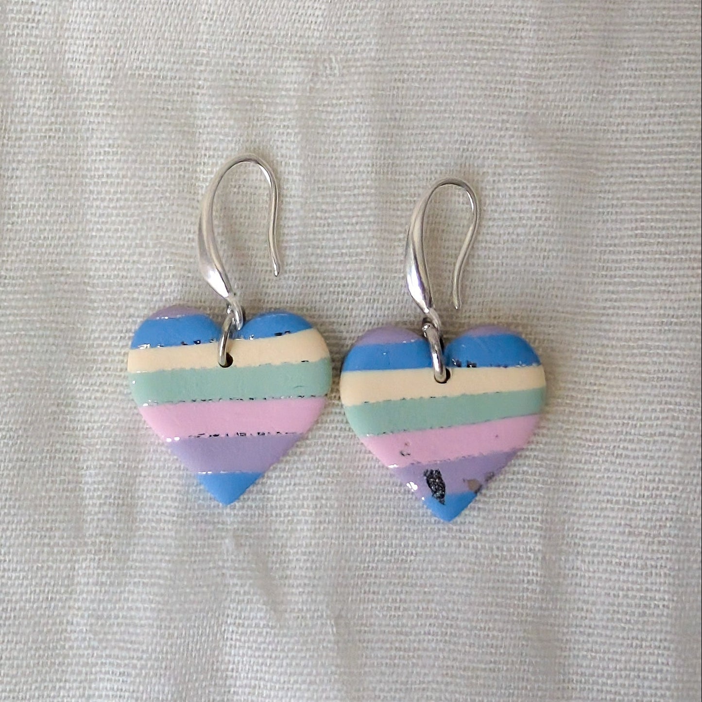 A pair of heart-shaped dangling earrings with horizontal pastel stripes in blue, cream, pink, and lavender, hanging from silver fishhook wires.