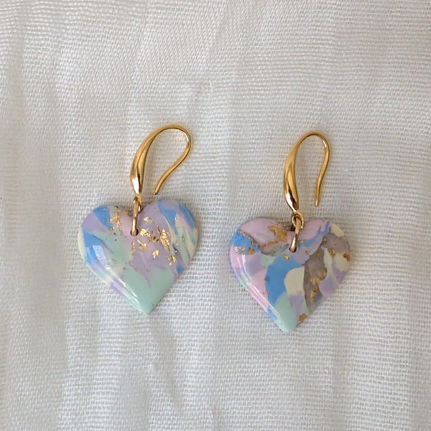A pair of heart-shaped drop earrings with a pastel marbled pattern in blue, pink, and mint green, accented with gold foil flakes and suspended from gold fishhook ear wires.