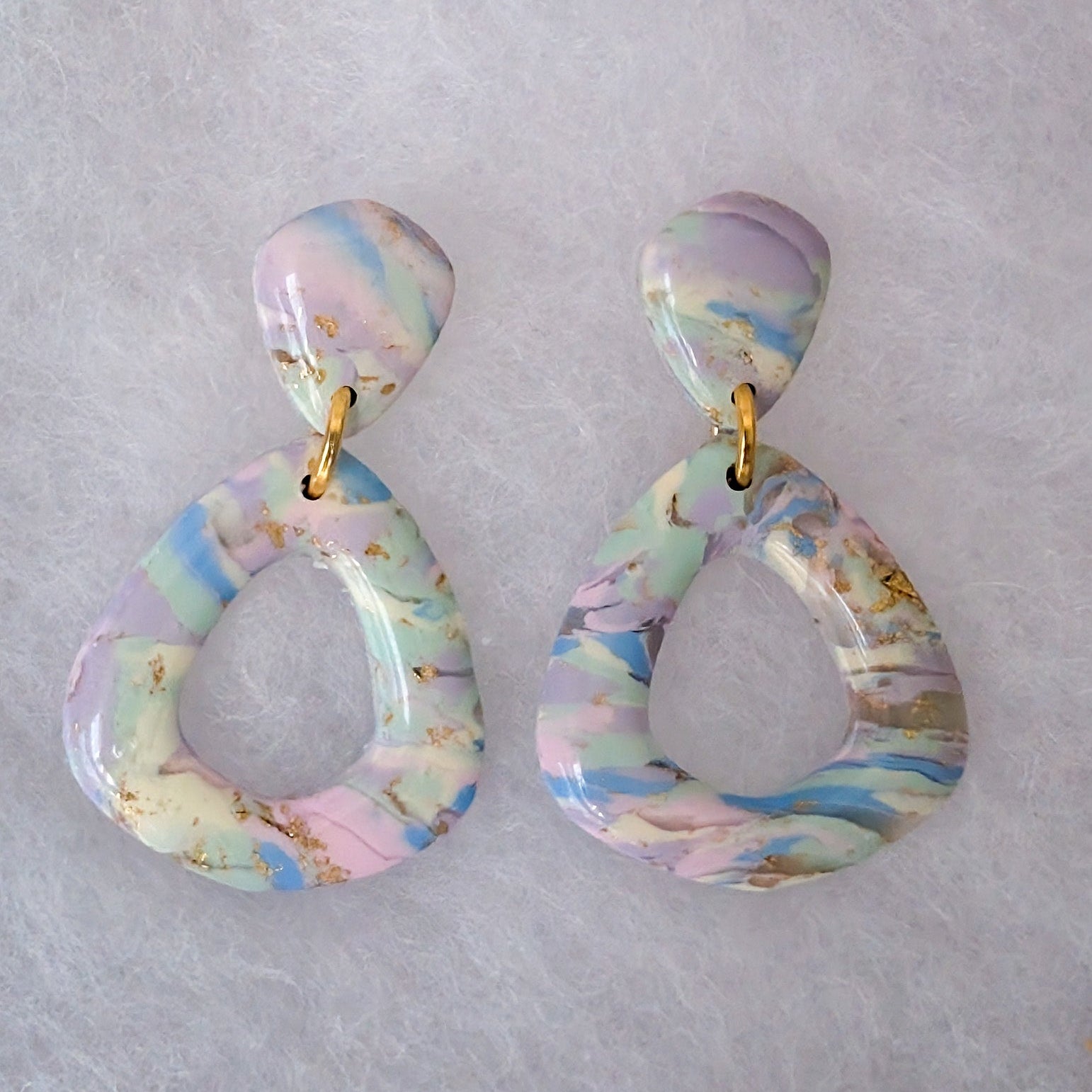 A pair of dangling teardrop-shaped earrings with a pastel marbled pattern in pink, blue, and purple, accented with gold flecks and connected by small gold rings.