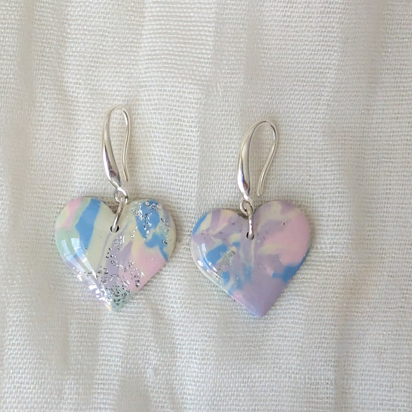 A pair of dangling heart-shaped earrings with a marbled pastel design in pink, blue, and lavender, suspended from silver fishhook wires.