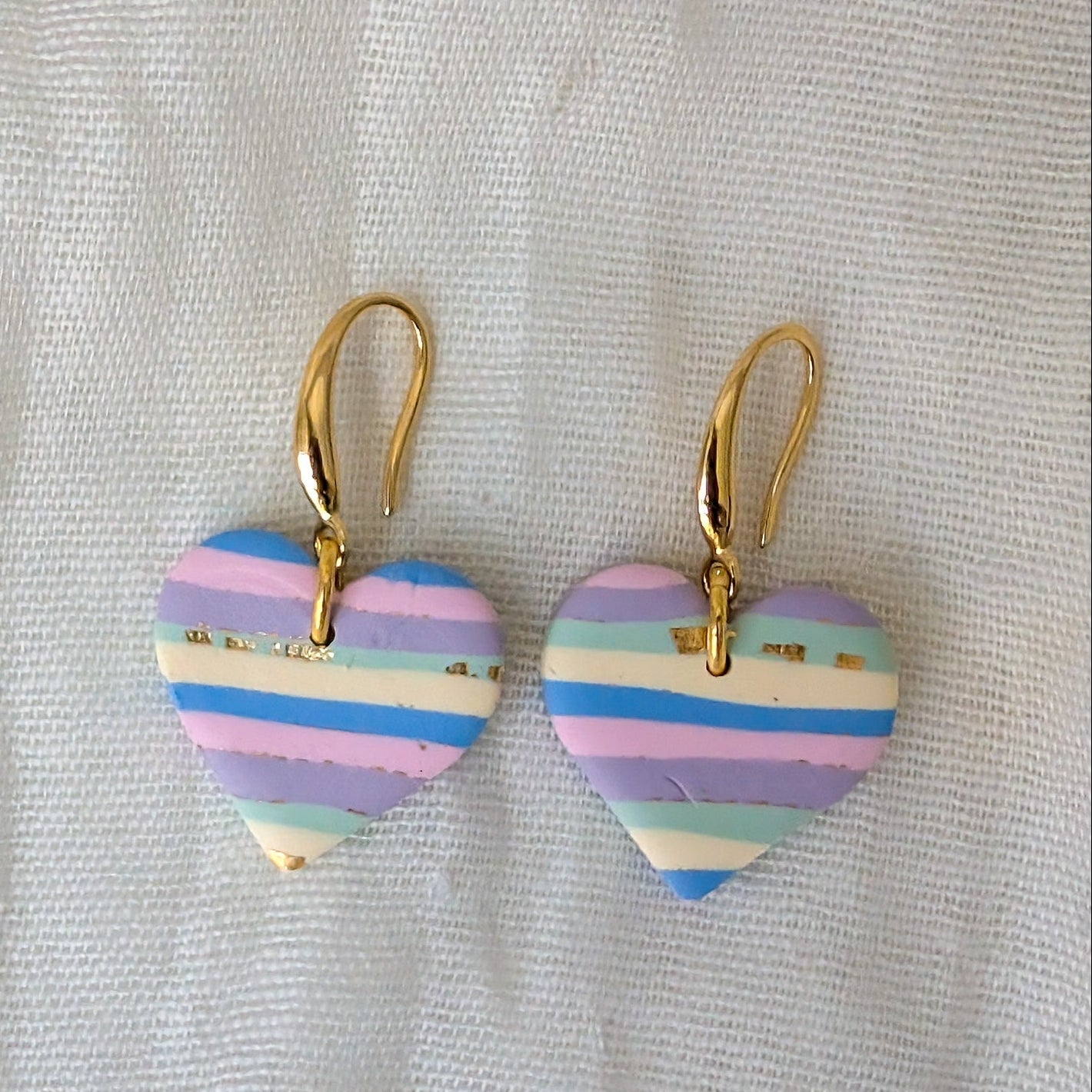 A pair of heart-shaped drop earrings with gold-tone fishhook ear wires, featuring pastel horizontal stripes in purple, blue, pink, and cream.