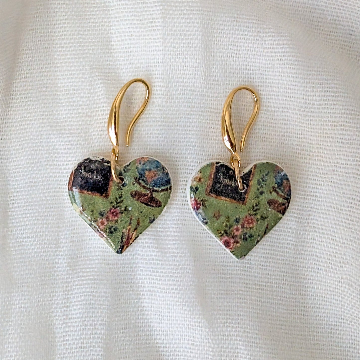 A pair of heart-shaped dangle earrings with gold-tone hooks and a vintage floral pattern in muted green, pink, and blue, displayed on a textured white fabric background.