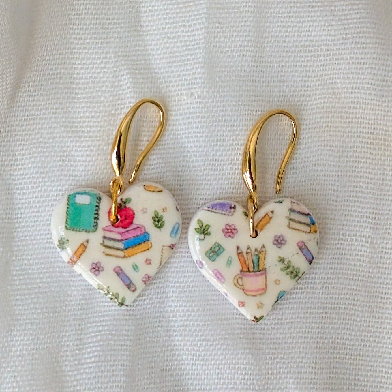 A pair of heart-shaped dangle earrings with gold hooks, featuring a white background decorated with colorful school-themed illustrations like books, pencils, and a cup.