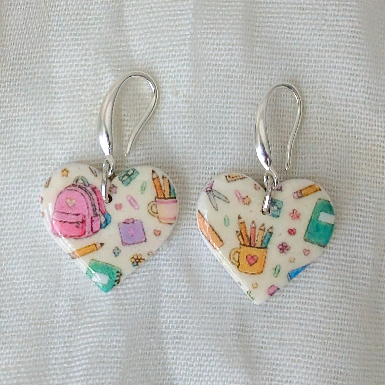 A pair of heart-shaped dangle earrings with a colorful school-themed pattern on a white background, hanging from silver fishhook wires.