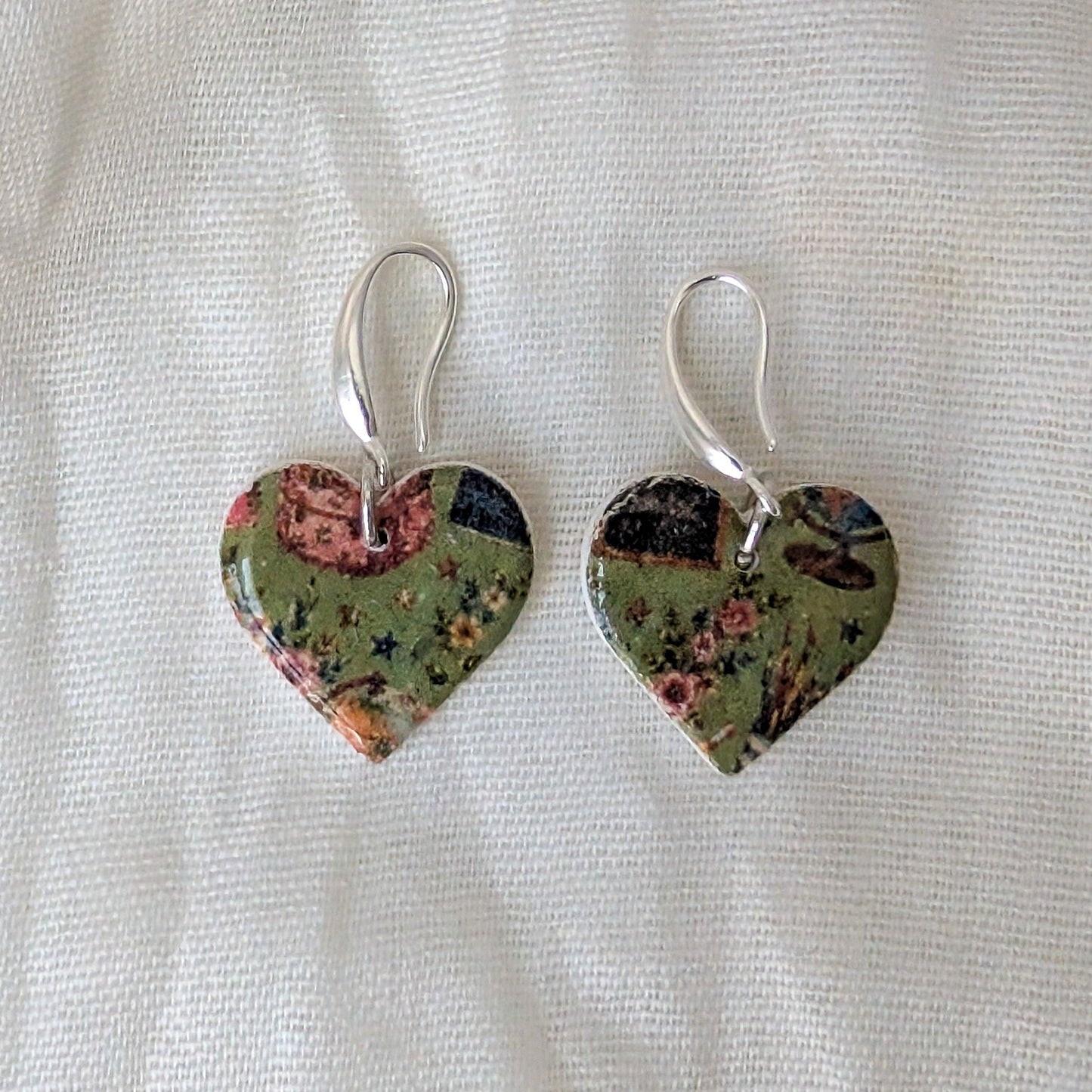 A pair of heart-shaped dangle earrings with a green floral and abstract patterned face, suspended from silver fishhook wires, displayed on a textured white fabric background.