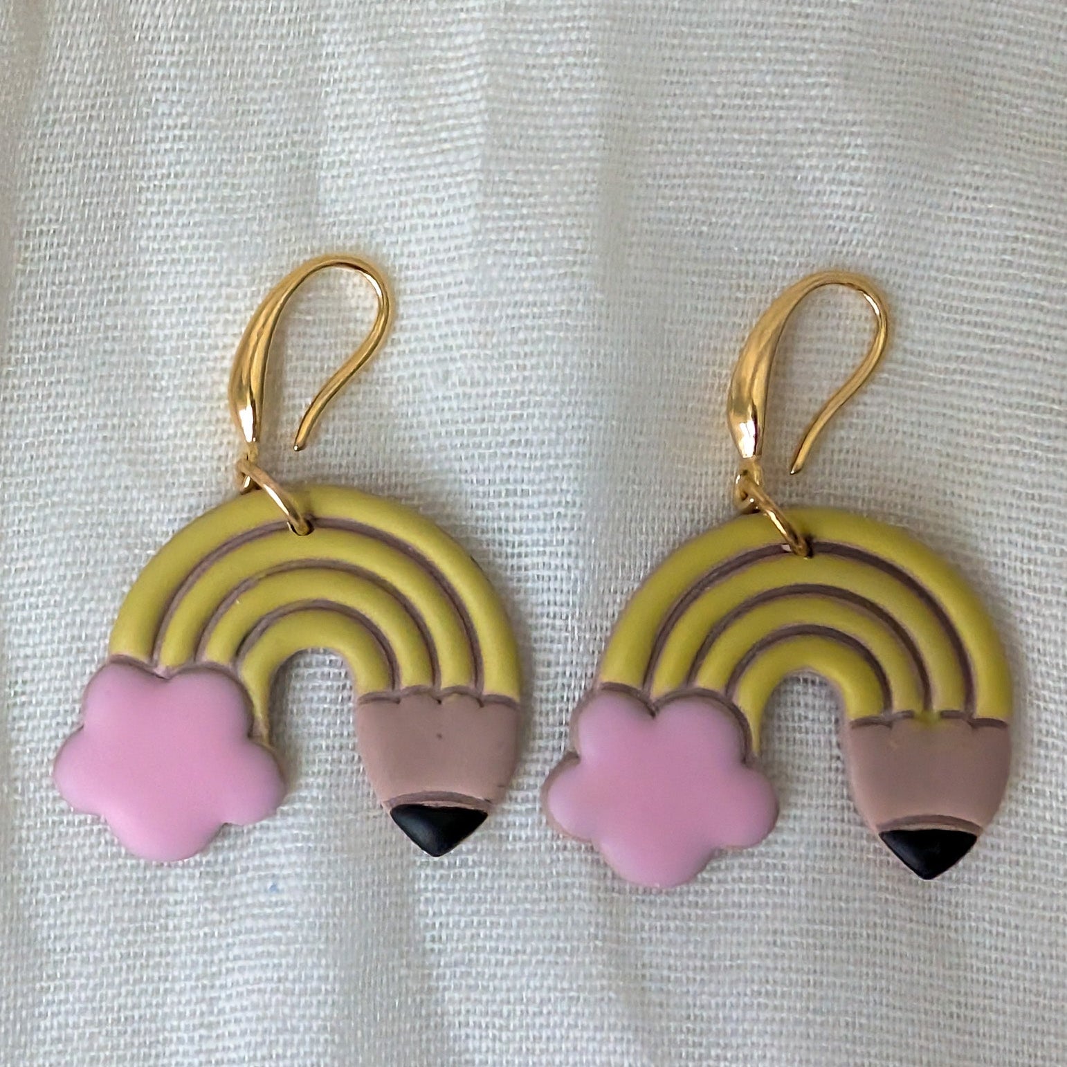A pair of gold-tone and pastel-colored dangling earrings shaped like mini rainbows with pink clouds and pencil tips, displayed on a textured white fabric background.