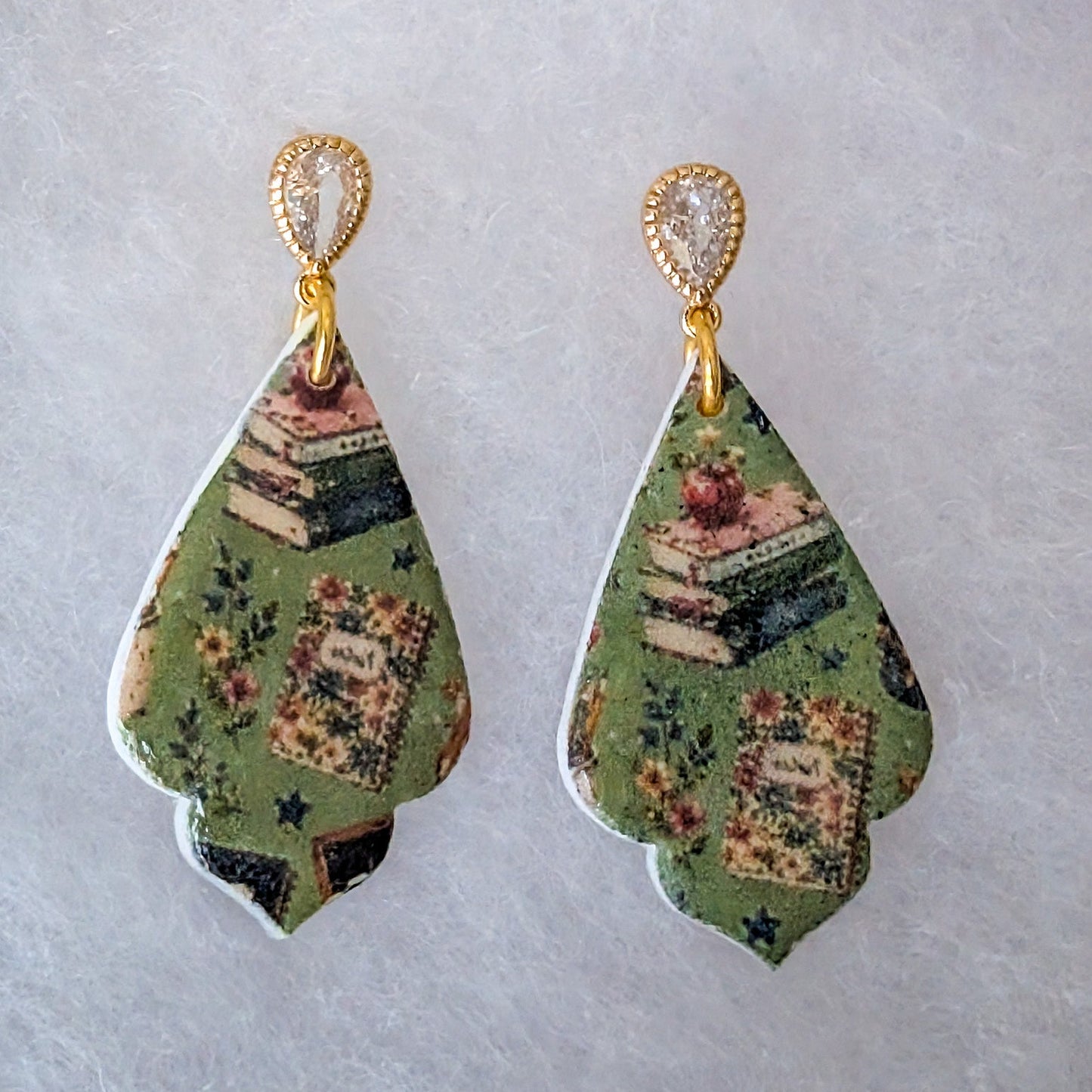 A pair of teardrop-shaped dangle earrings with a vintage book and floral pattern on a sage green background, featuring gold-tone posts and clear rhinestone accents, displayed on a textured light gray surface.