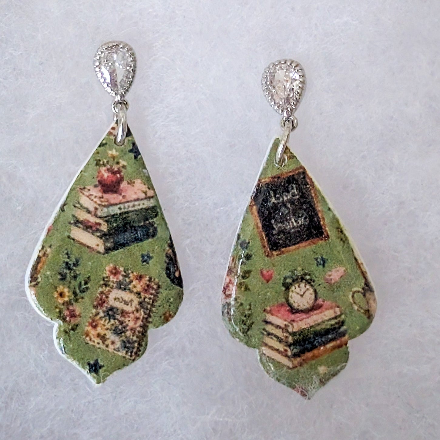 A pair of handmade teardrop-shaped earrings made from green patterned paper featuring vintage book illustrations, with silver-toned metal studs, displayed on a light gray textured background.
