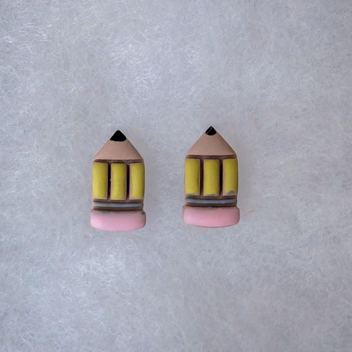 A pair of pencil-shaped stud earrings on a textured light gray background. The earrings feature a light pink base, yellow pencil bodies.