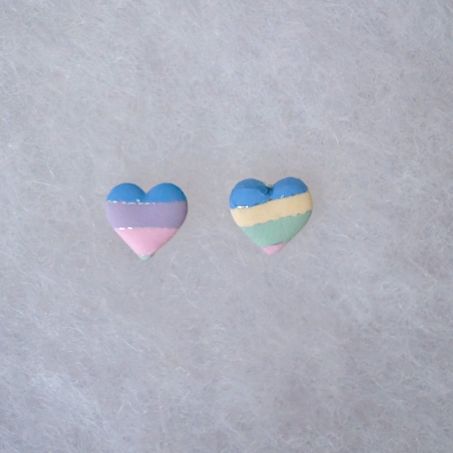 Two small heart-shaped stud earrings with horizontal pastel stripes in blue, lavender, pink on one earring and blue, cream, mint, and pink on the other with silver accents.