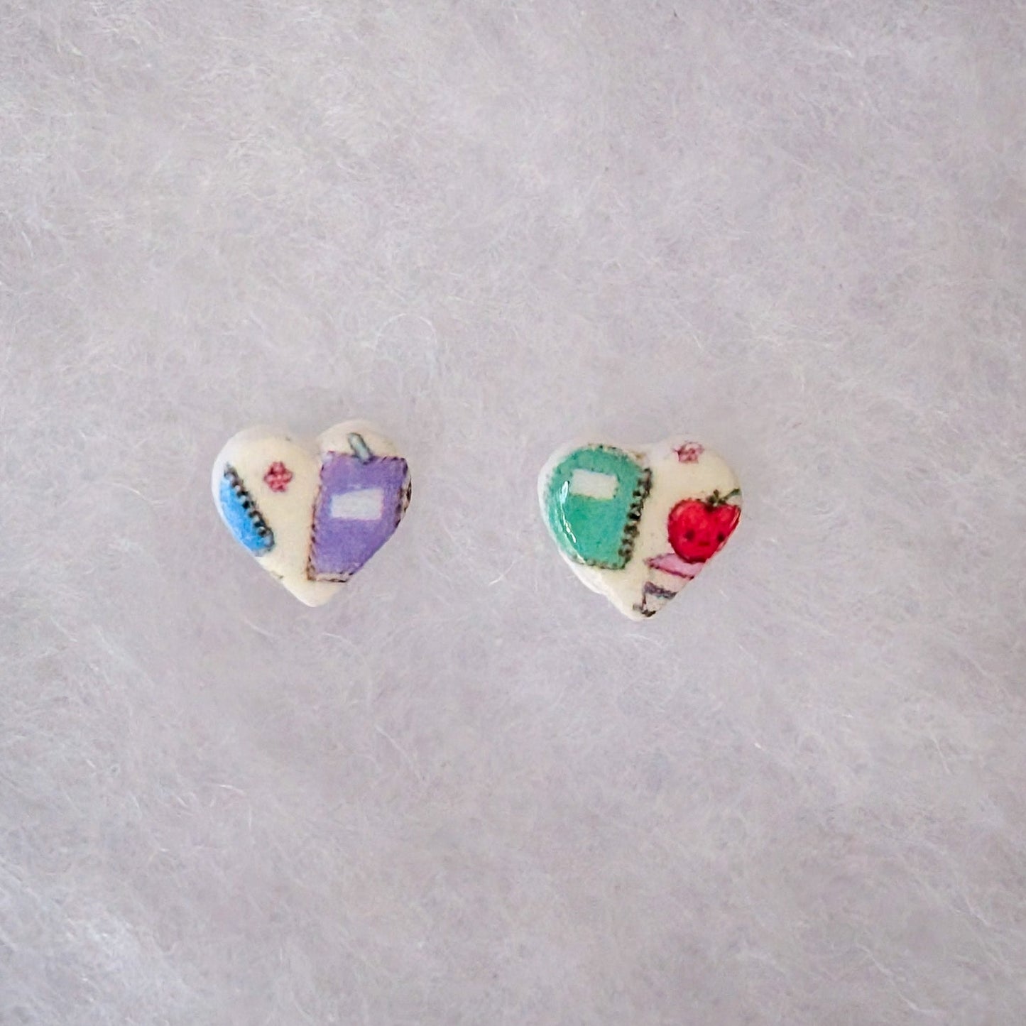 A pair of small heart-shaped stud earrings. One earring features a purple post with a white heart decorated with a blue and red striped gem and a silver heart.
