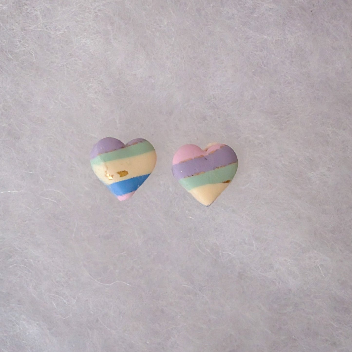 Two small heart-shaped earrings with pastel rainbow stripes in purple, blue, green, yellow, and pink with gold accents.