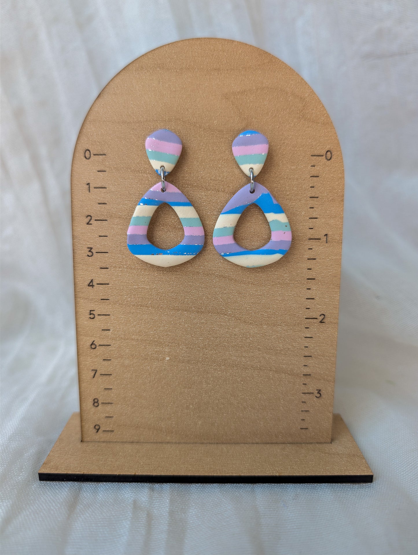 A pair of dangling earrings displayed on a wooden arch stand with measurement markings. The earrings have pastel horizontal stripes in pink, blue, purple, and cream, with a small silver-tone ring connector at the top.