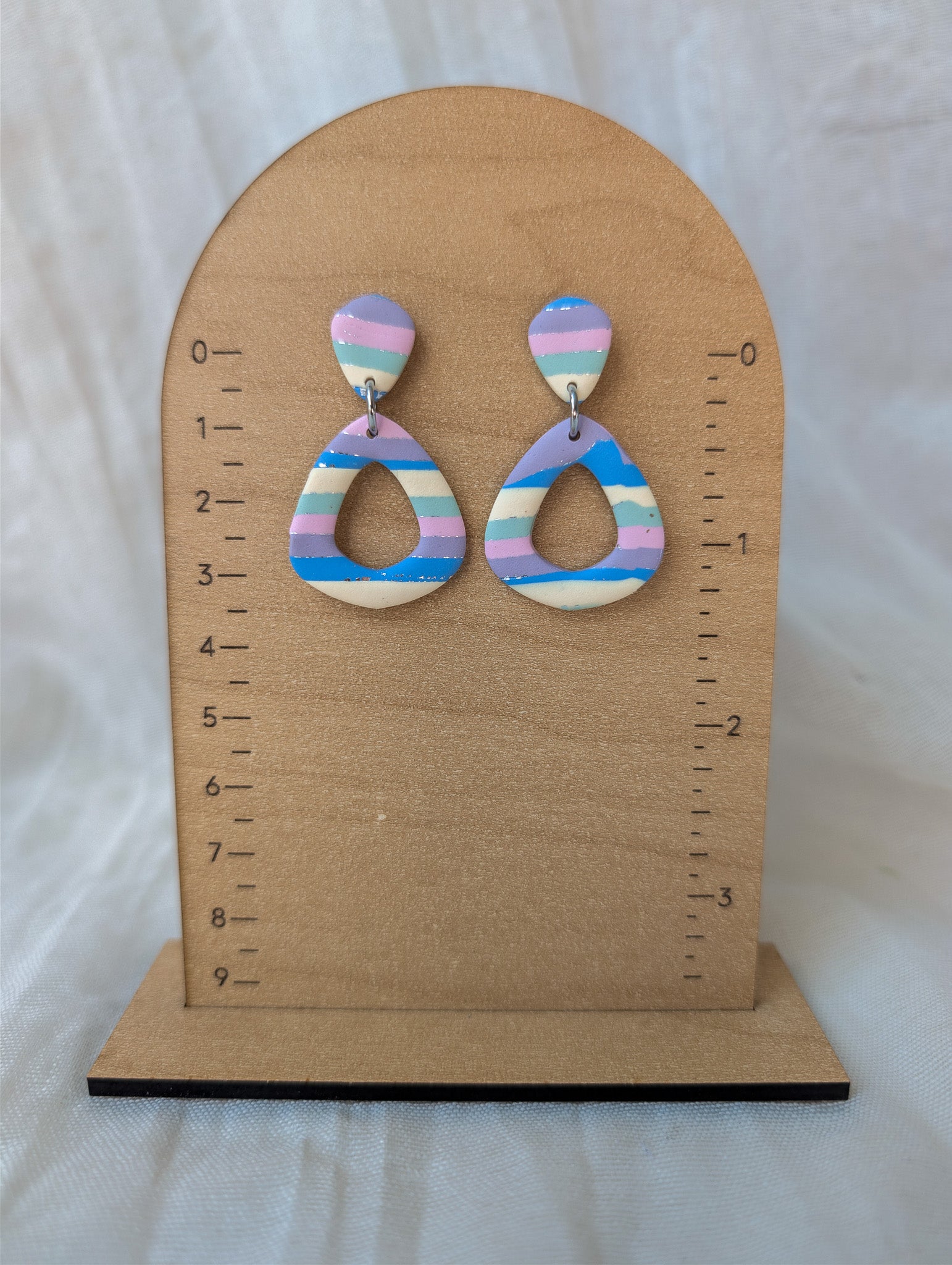 A pair of dangling earrings displayed on a wooden arch stand with measurement markings. The earrings have pastel horizontal stripes in pink, blue, purple, and cream, with a small silver-tone ring connector at the top.