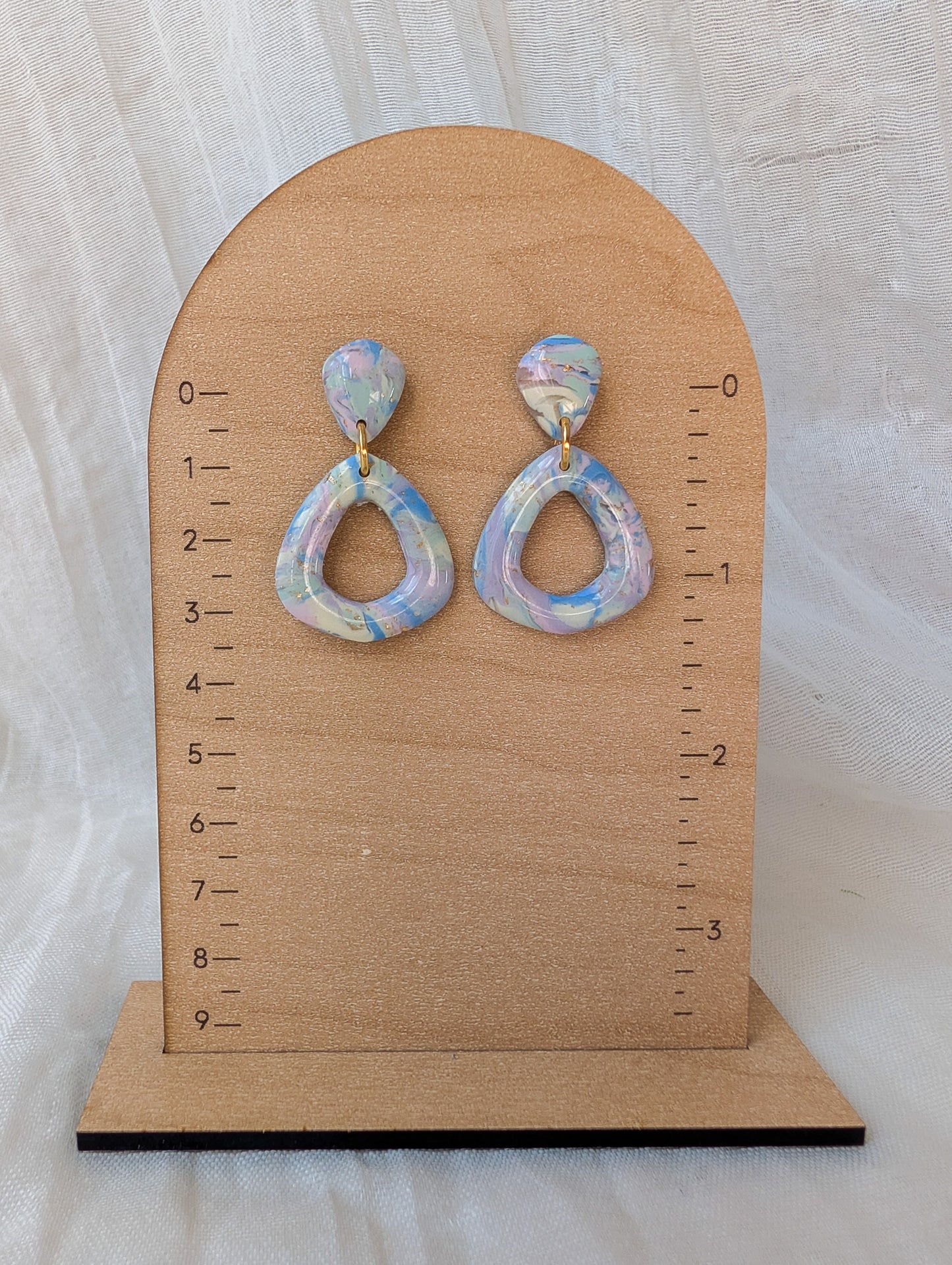 A pair of dangling teardrop-shaped earrings with iridescent pastel blue, white, and lavender opal-like centers, displayed on a wooden arch stand with measurement markings.