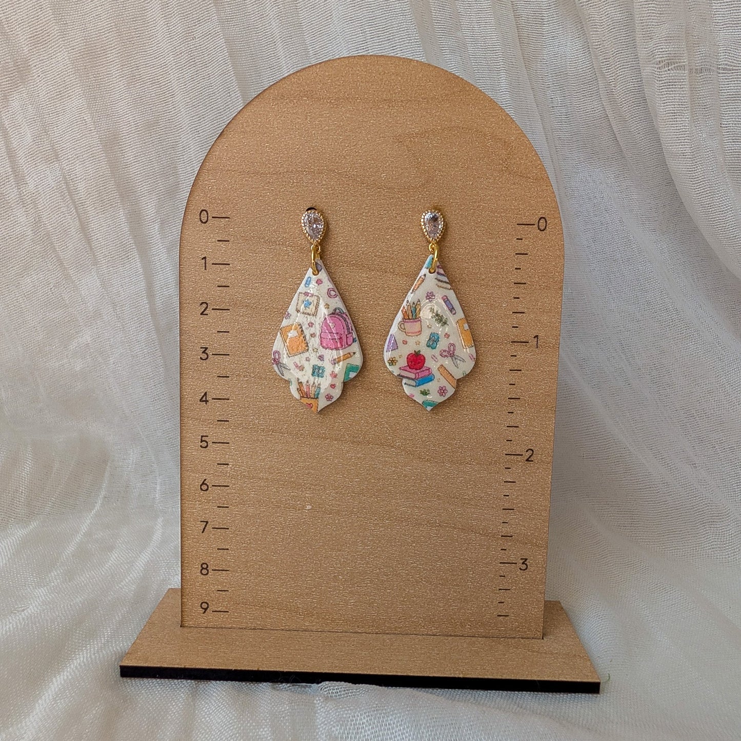 A pair of teardrop-shaped dangle earrings with a colorful, patterned fabric design, displayed on a wooden arch stand with measurement markings.