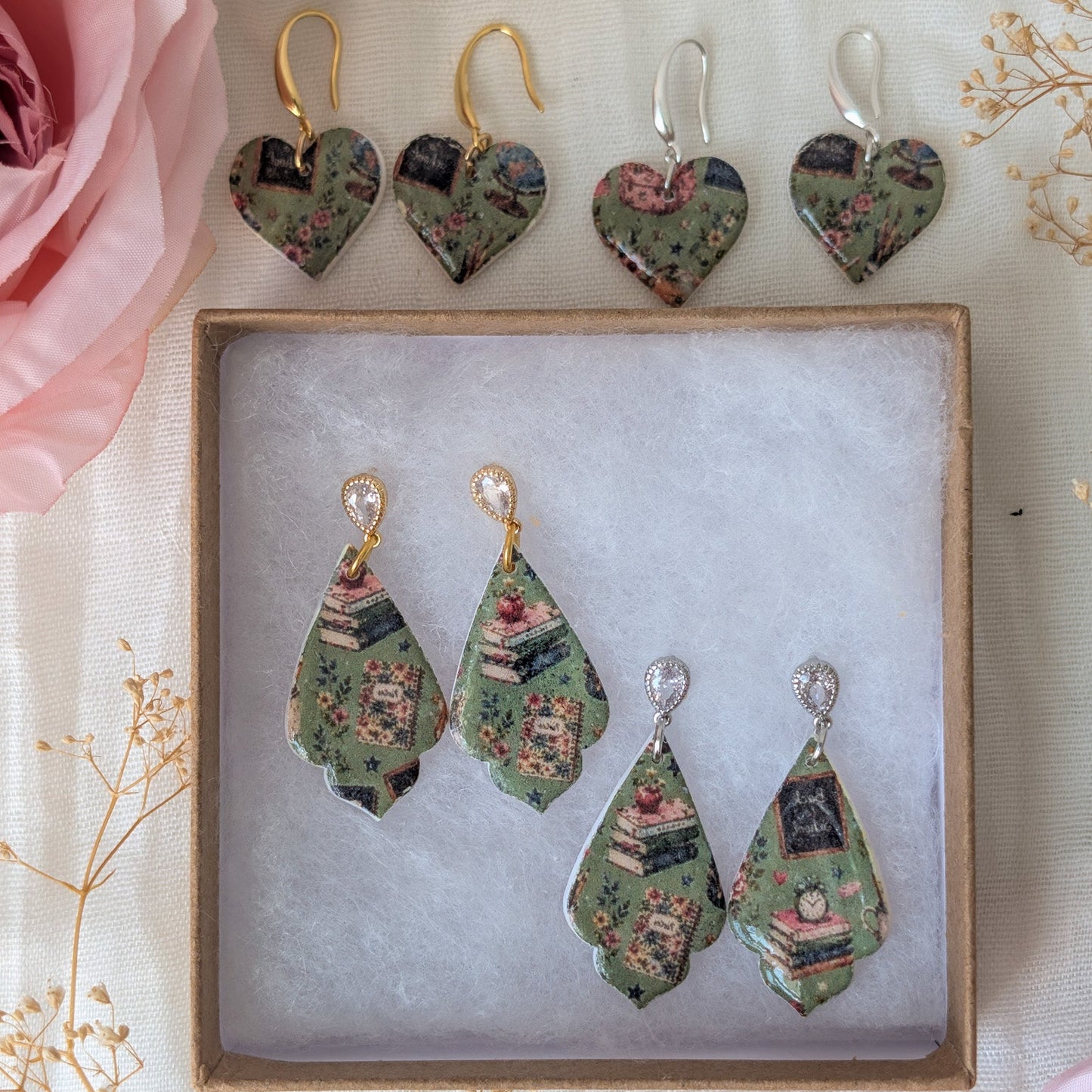 Four pairs of handmade earrings displayed in a square wooden box on a light fabric background, with pink roses and dried flowers as props. The earrings feature a vintage book collage pattern in muted green, pink, and brown tones, with gold and silver hooks.