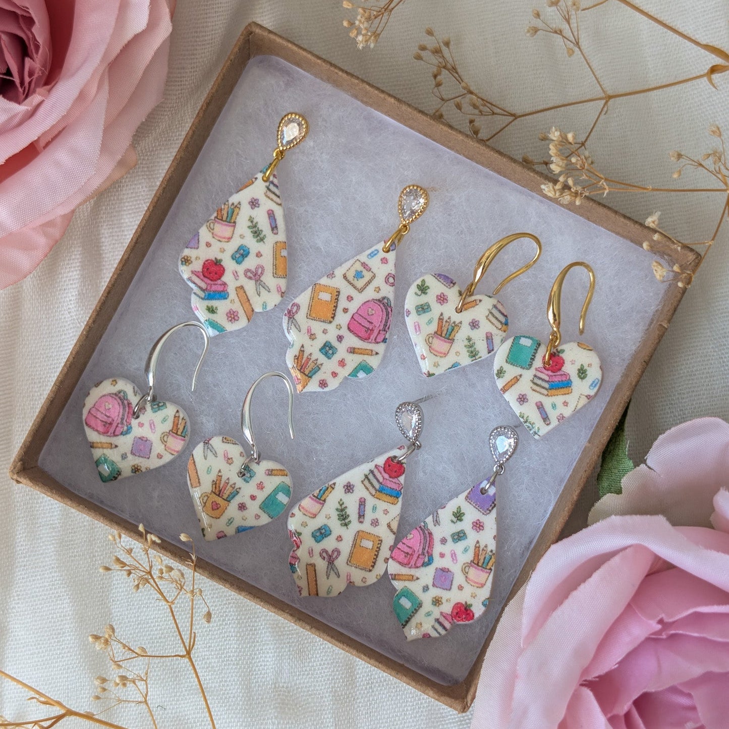 A set of heart-shaped earrings with a colorful cupcake print, displayed in a brown gift box on a light fabric background with pink roses and dried flowers.