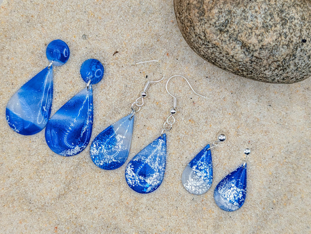 Blue teardrop earrings with silver hooks on a sandy surface