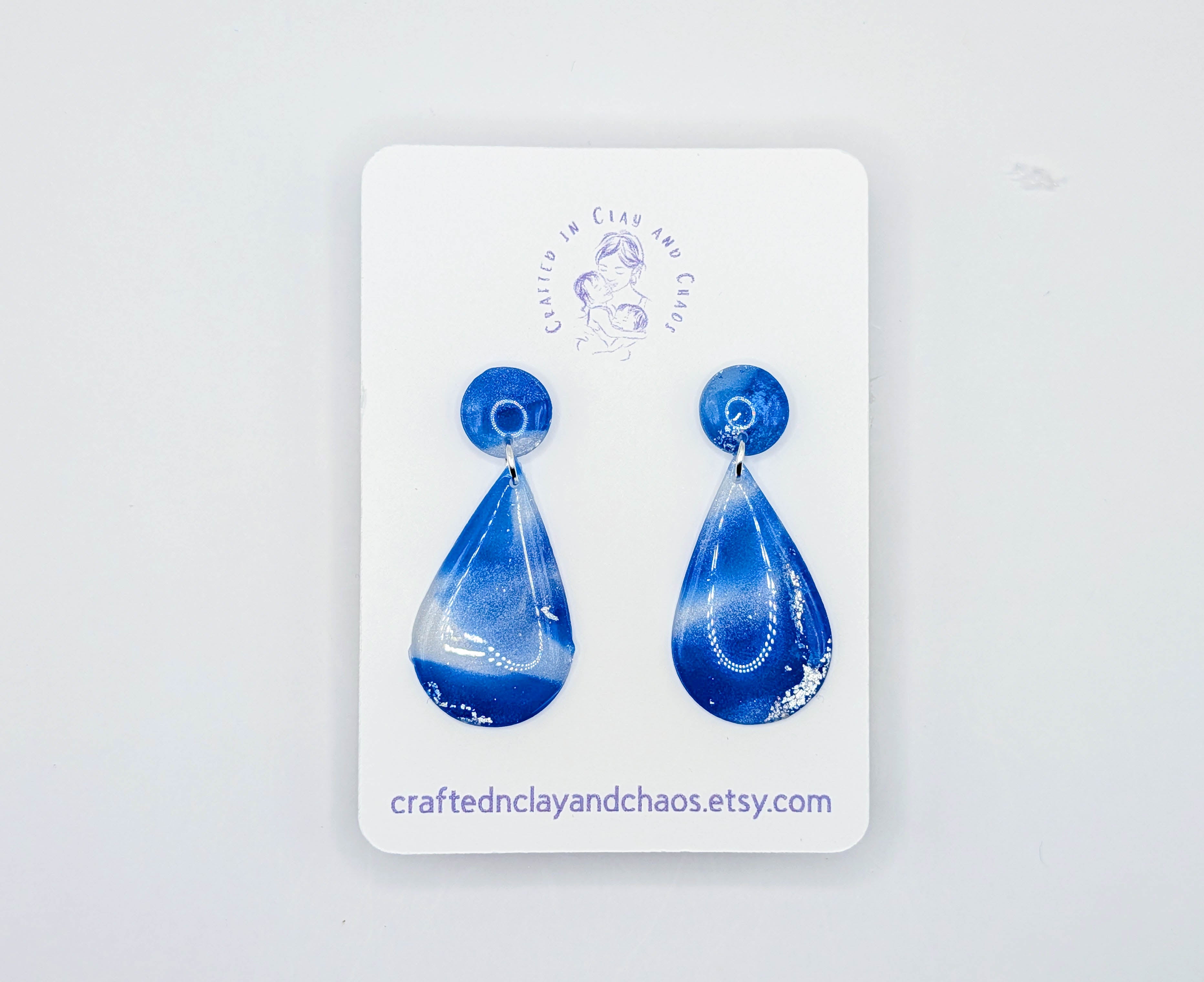 Blue teardrop earrings on a white card with 'Crafted in Clay and Chaos' branding.