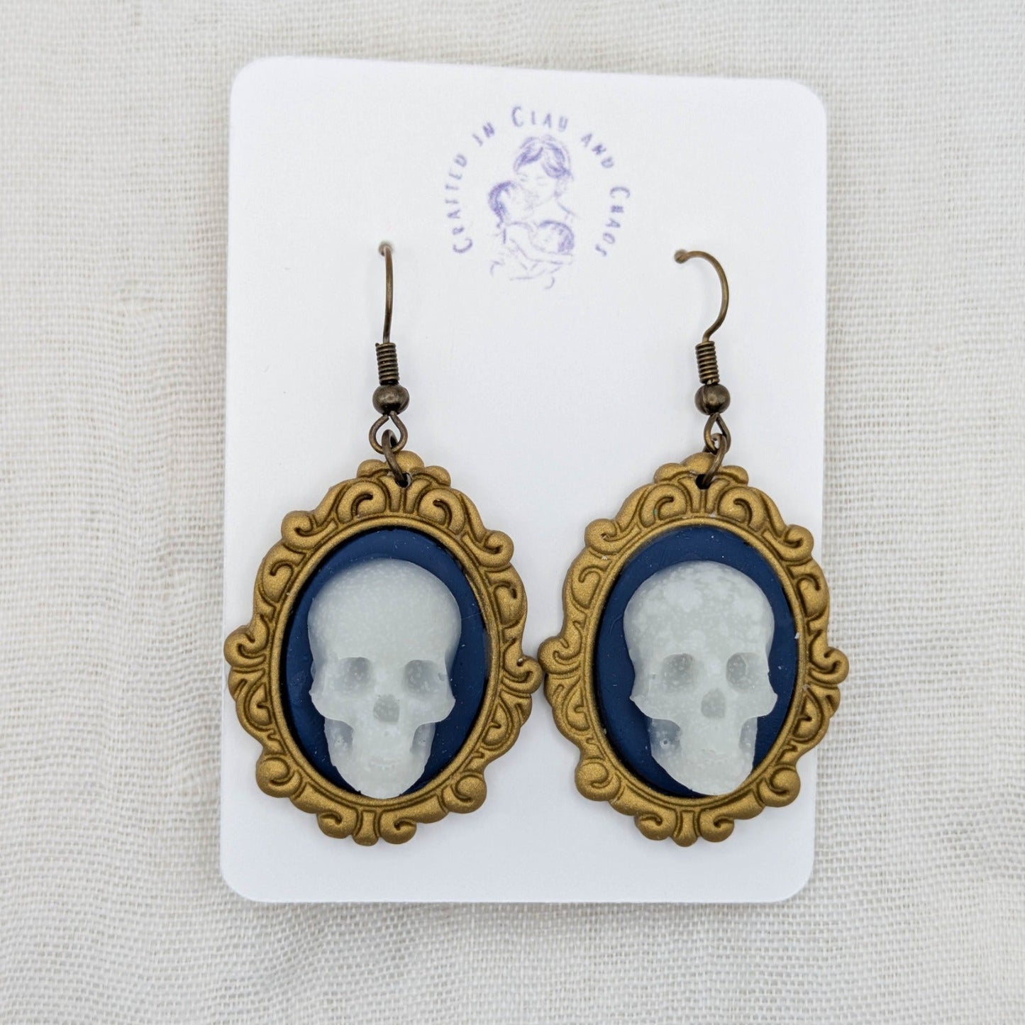 Second one-of-a-kind antique gold oval frame pair with white glow-in-the-dark skulls, blue background, and bronze-tone hooks