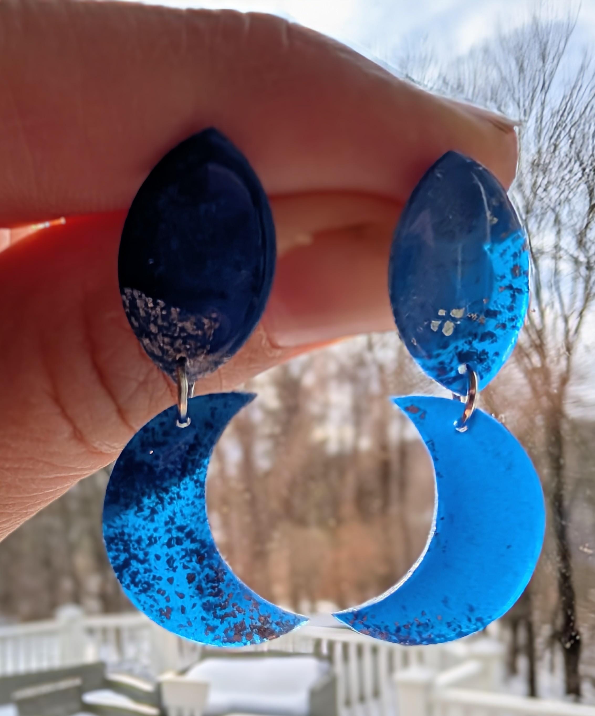Blue crescent moon earrings held in front of a snowy landscape.